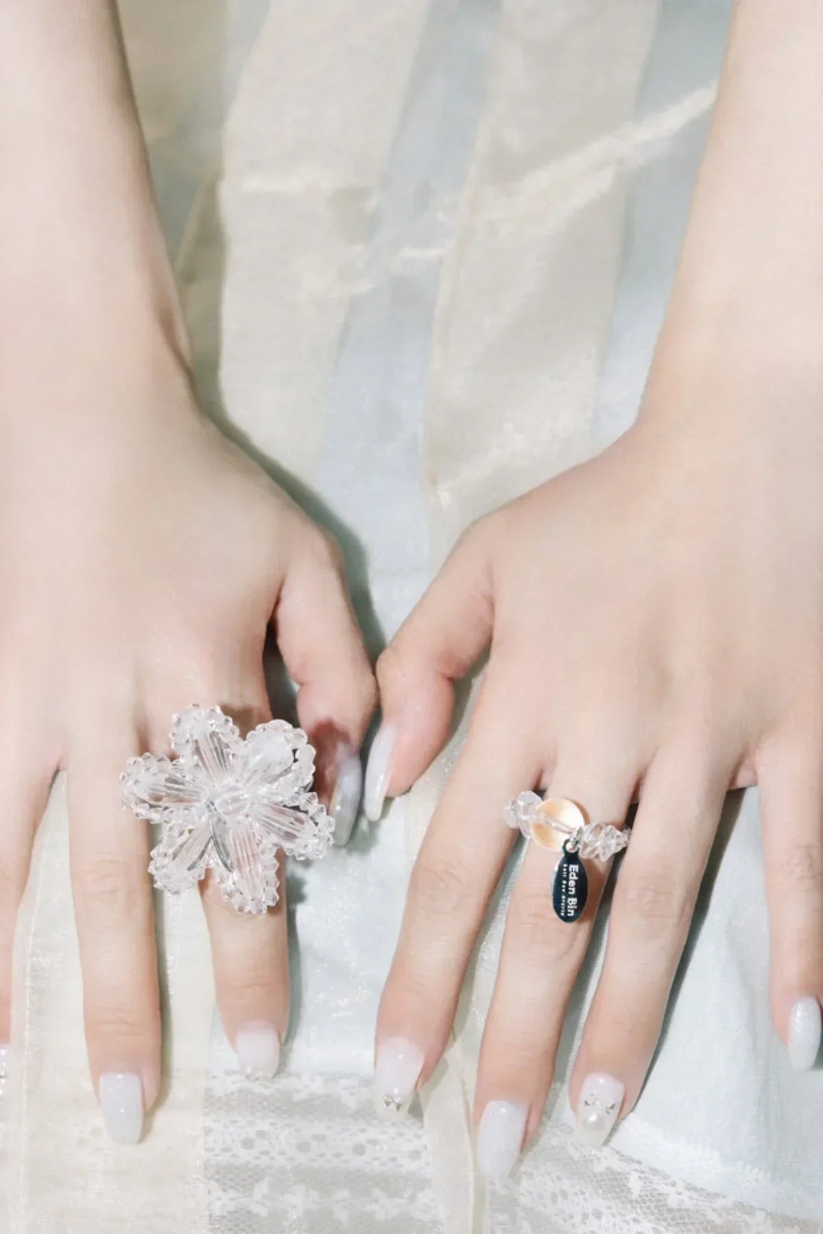 Close-up of hands wearing two clear resin rings with decorative nails on a lace fabric background