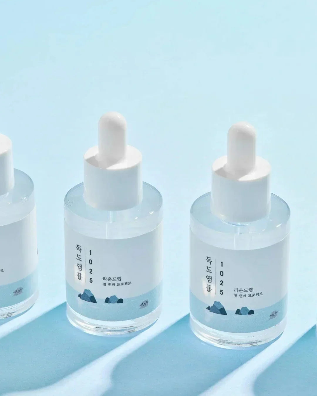 Three clear glass ampule bottles with white droppers on light blue background, labeled 1025 Dokdo Ampule skincare