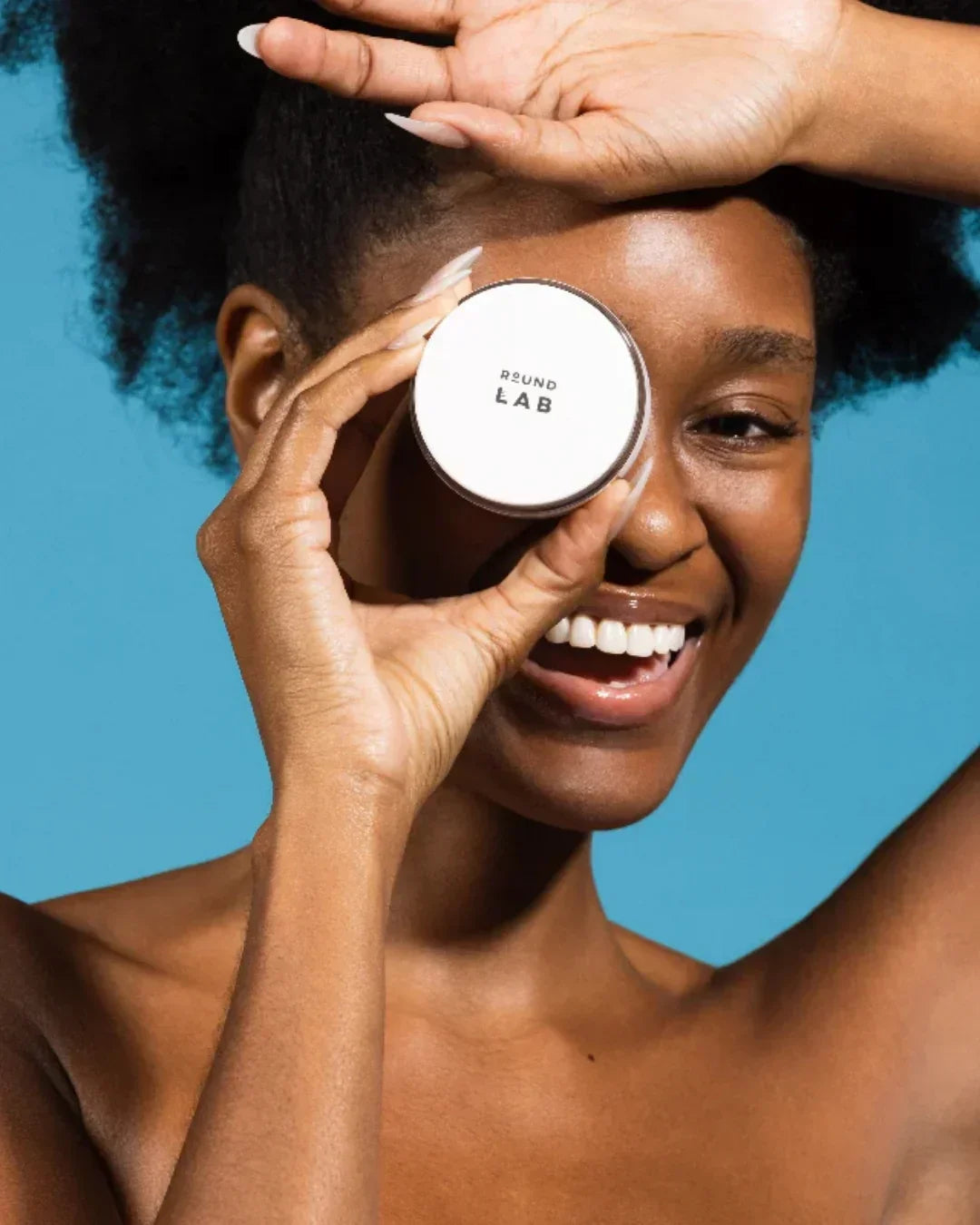 Smiling woman holding Round Lab skincare cream container over one eye against blue background