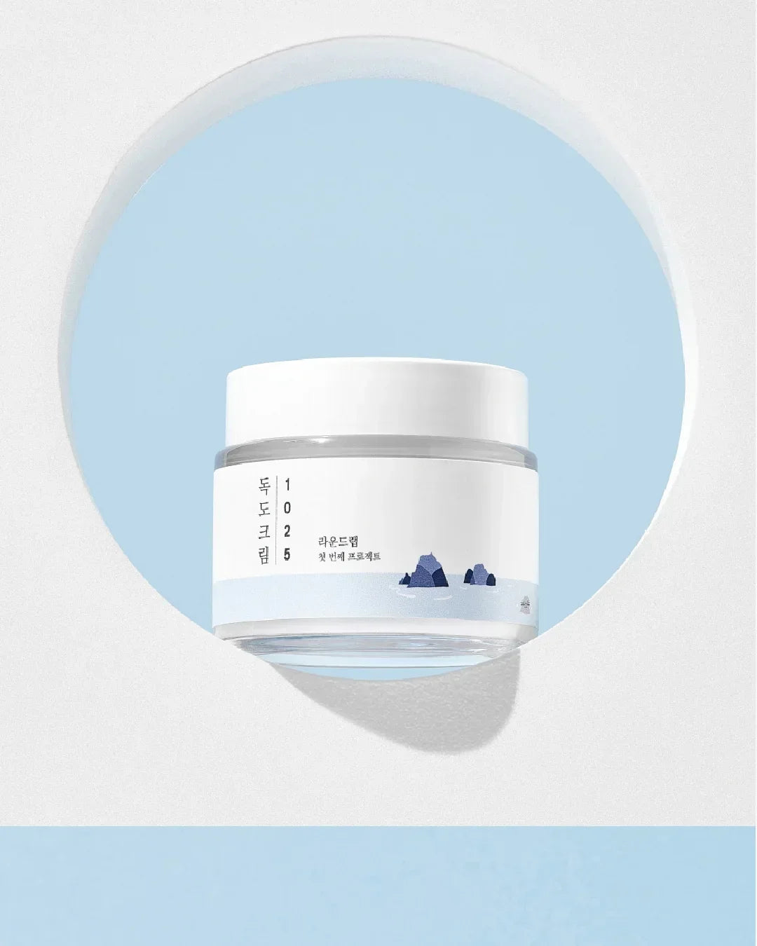 1025 Dokdo Cream in white jar with blue rock illustration on label, set against blue and white circular backdrop