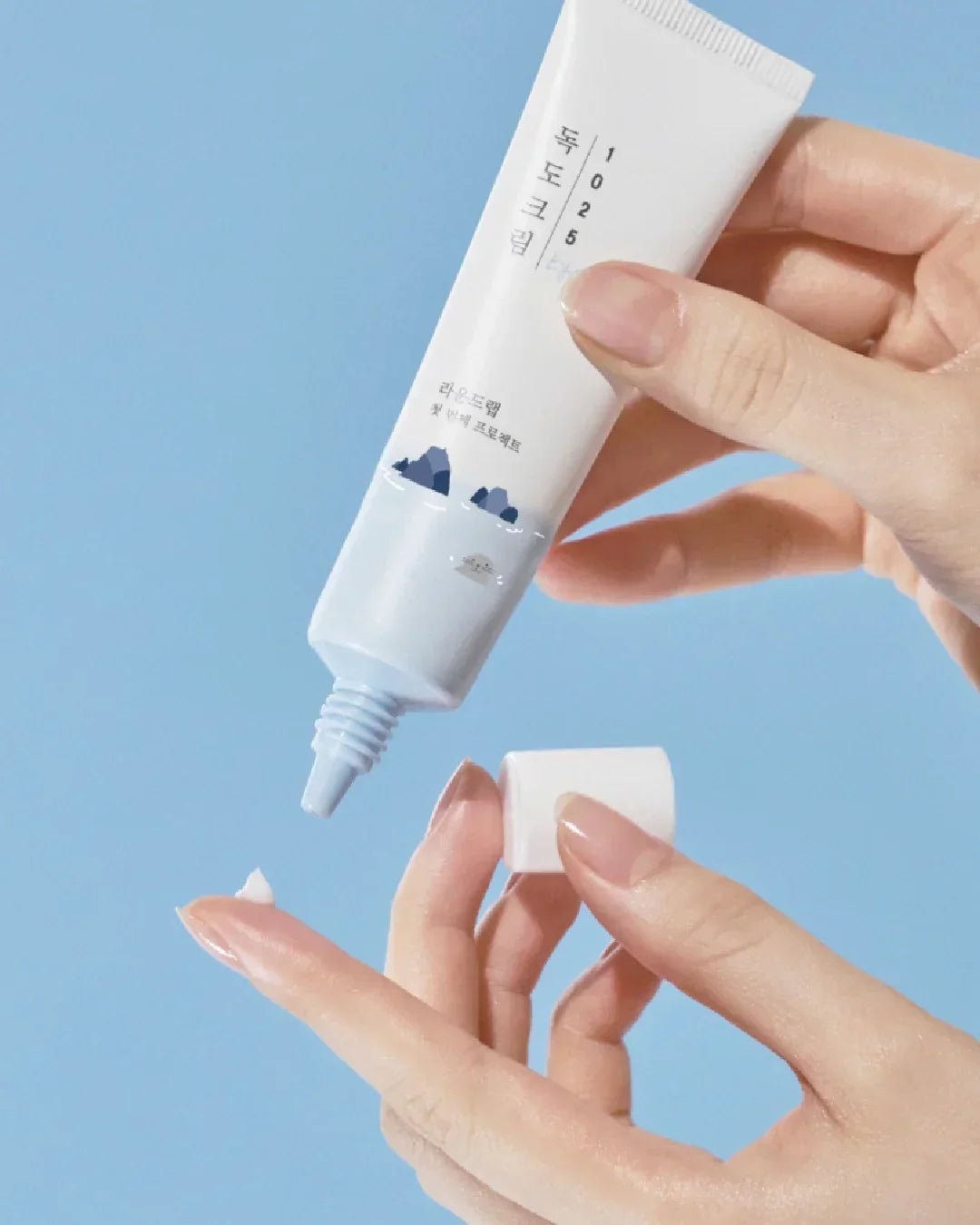 Hand holding and applying Dokdo 1025 eye cream from white tube with blue mountain design against light blue background