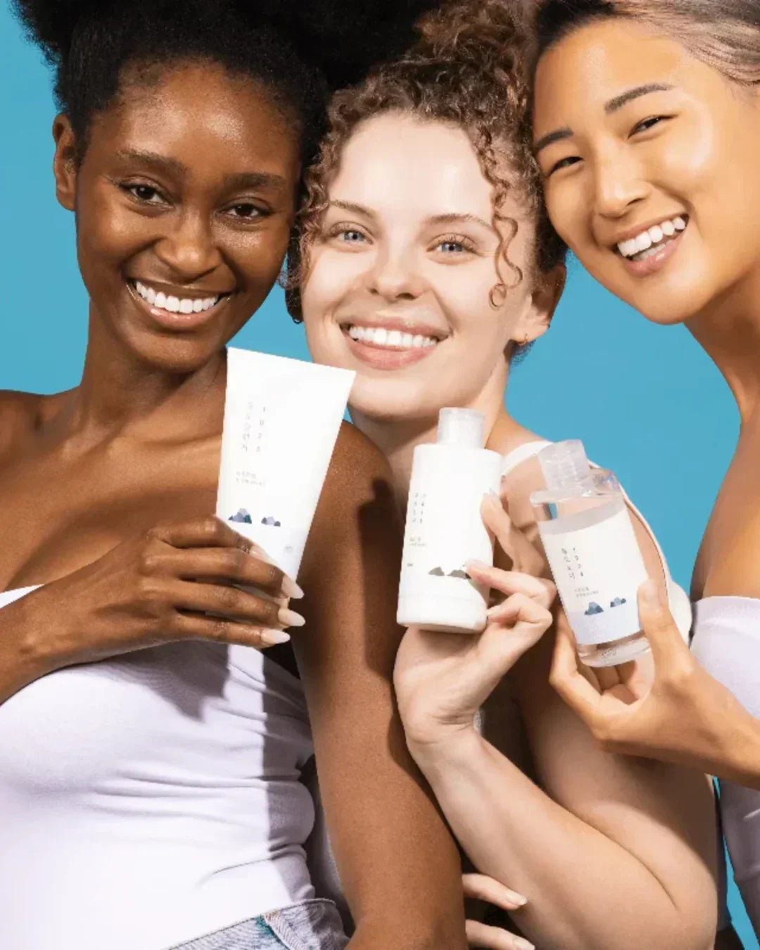 Three diverse women smiling and holding white skincare product bottles against blue background