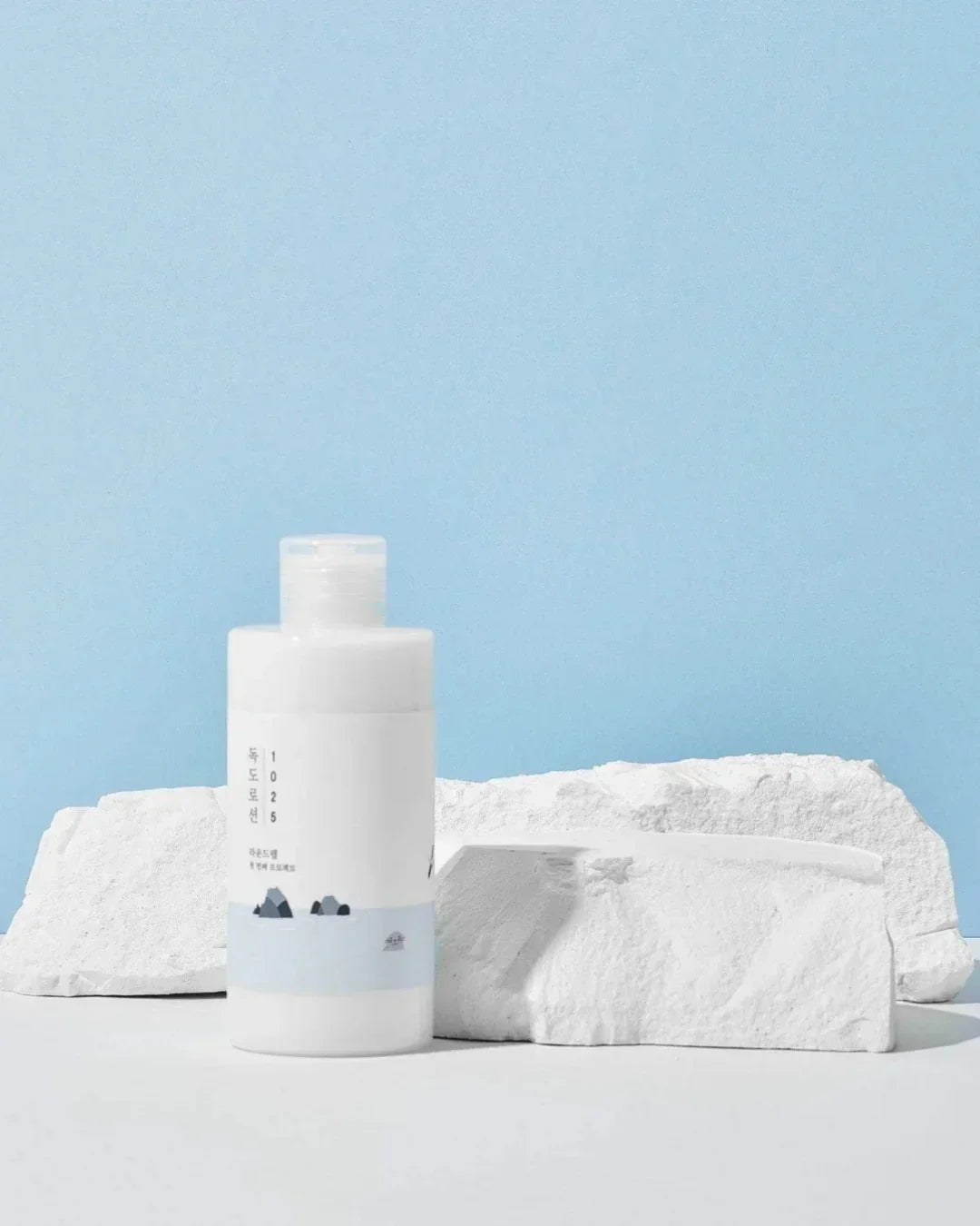 White Dokdo 1025 lotion bottle with Korean text, placed on white textured stones against light blue background