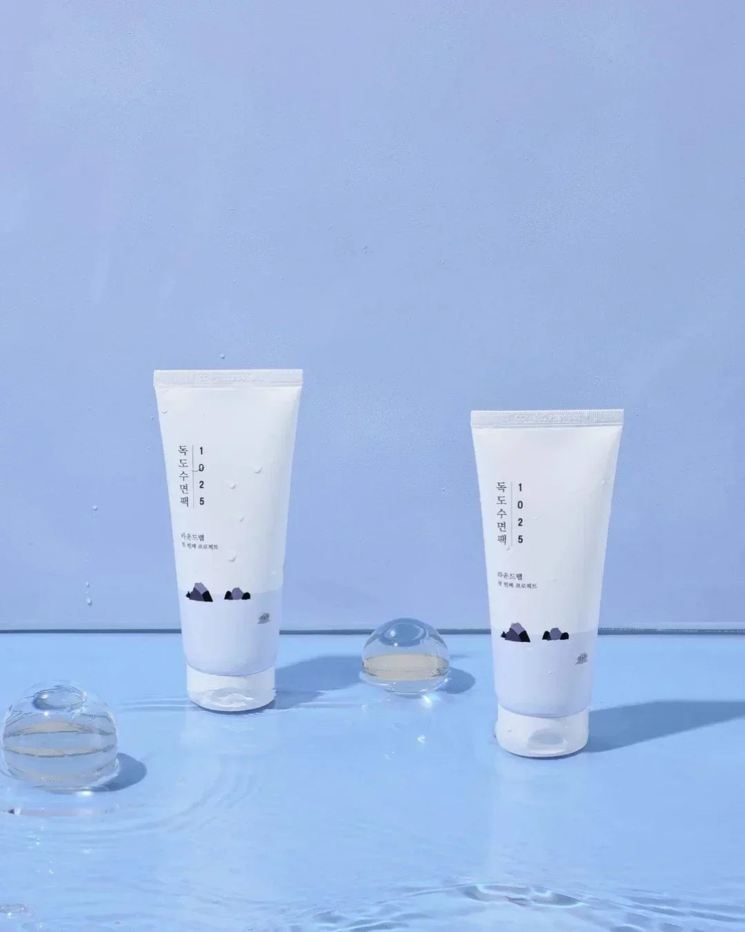 Two white tubes of skincare product Dokdo Sleeping Pack 1025 on blue surface with water droplets
