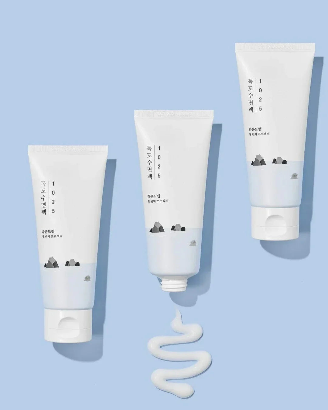 Three white tubes of 1025 Dokdo Sleeping Pack skincare product on light blue background with cream swirl