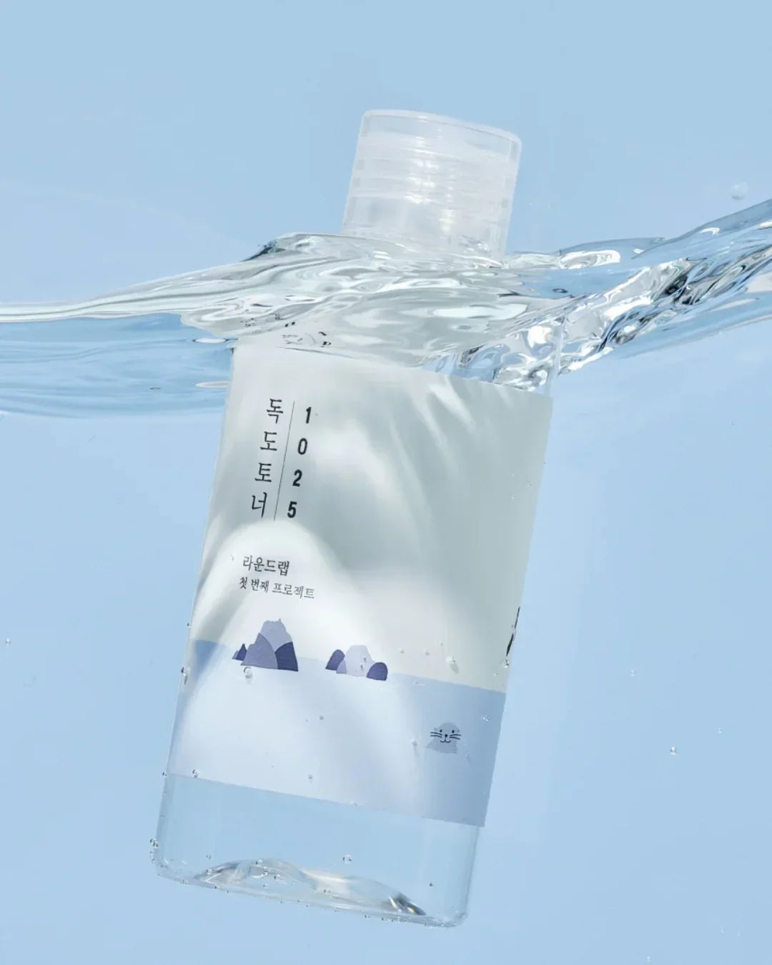 Clear plastic bottle labeled Dokdo Toner 1025 partially submerged in water with blue sky background