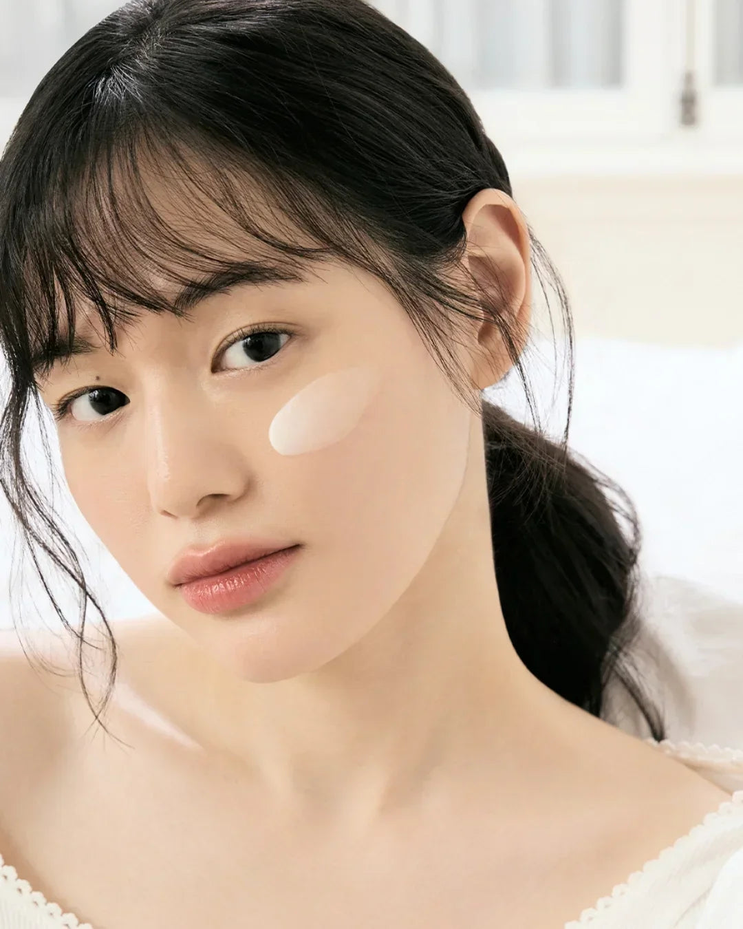 Close-up of a woman with smooth skin and a white cream swatch on her cheek, natural light
