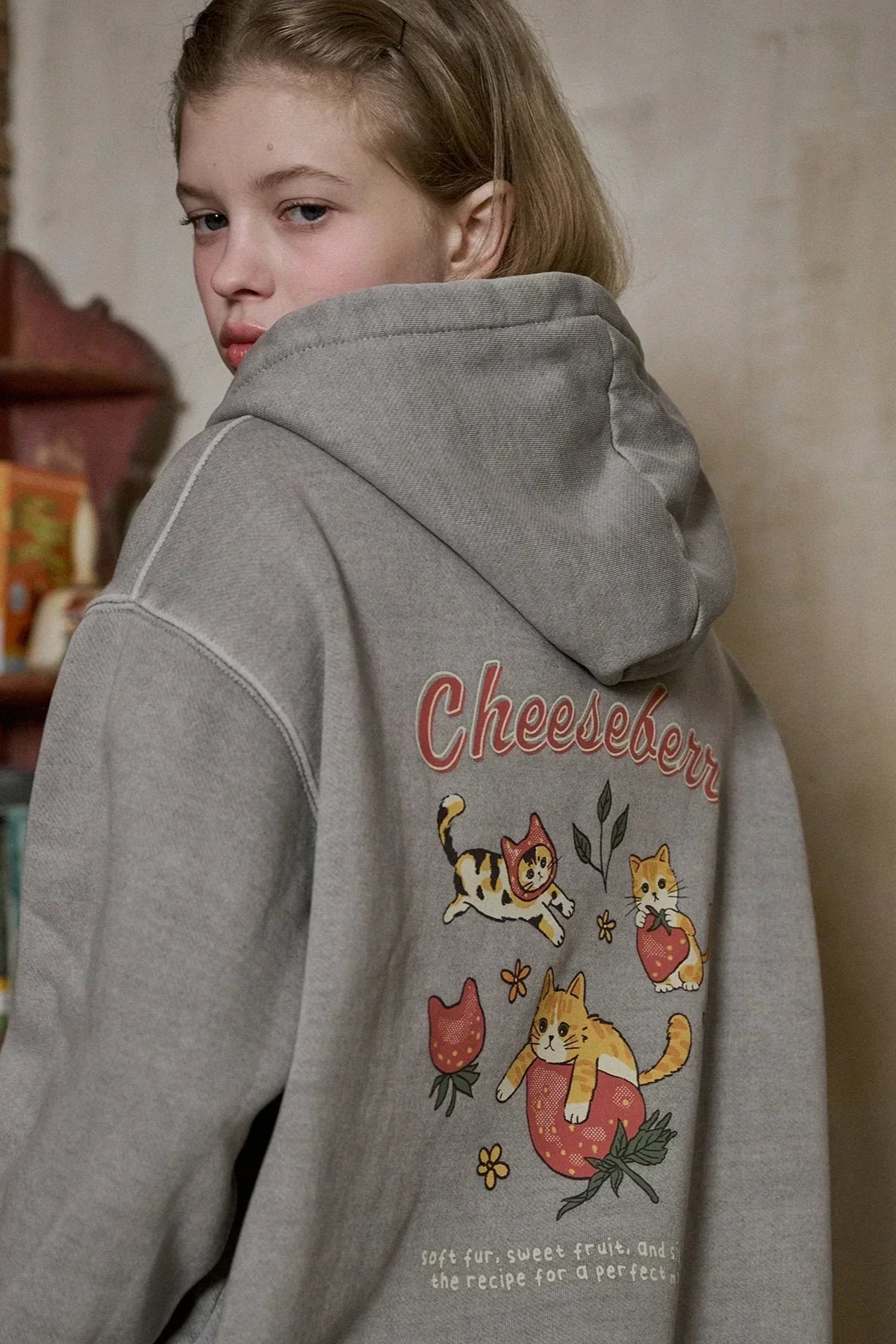 Young woman wearing gray hooded zip-up sweatshirt with playful cheeseberry cat and strawberry graphic on back