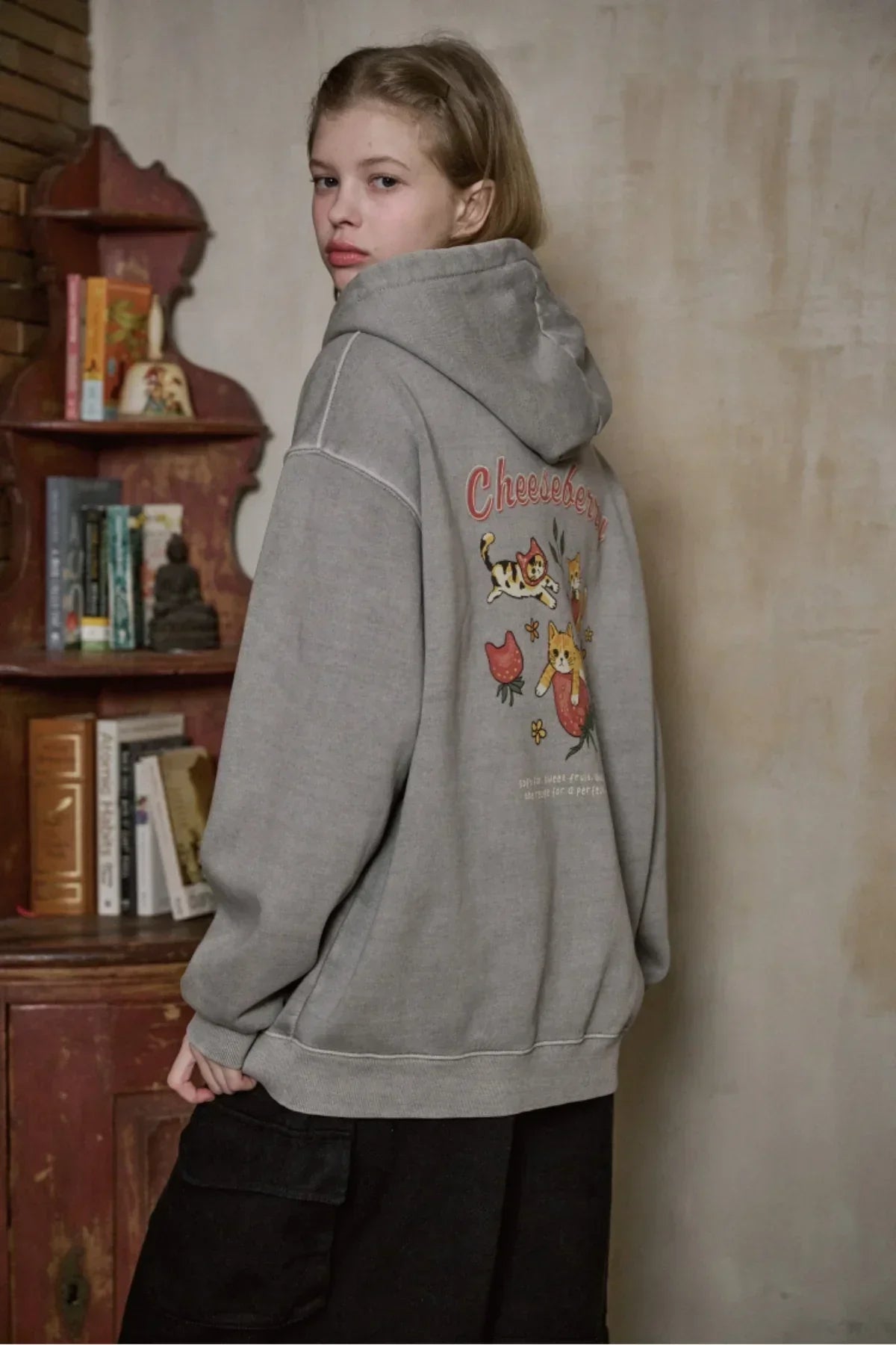 Young woman wearing ghost gray hooded zip-up sweatshirt with Cheesberry cat print, standing indoors