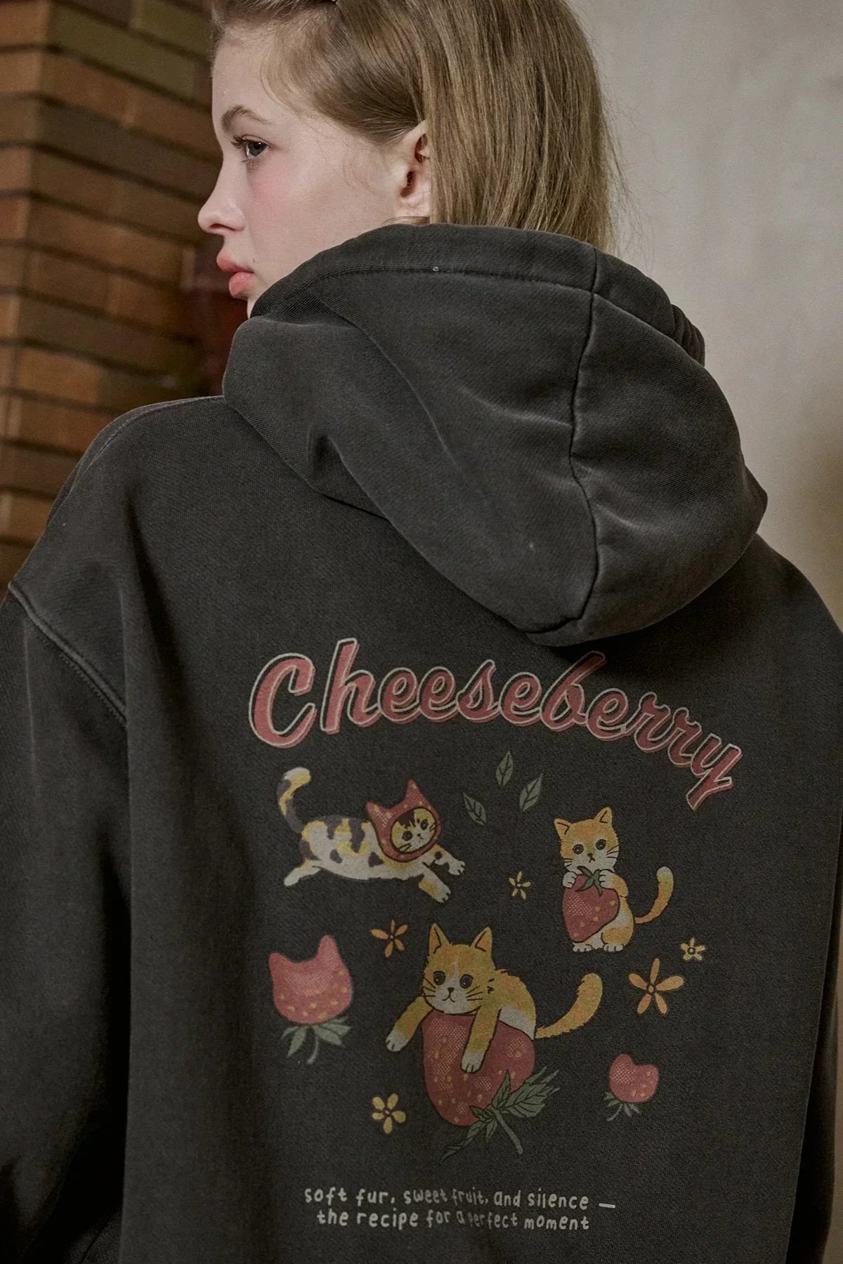 Close-up of a woman wearing a dark hooded zip-up sweatshirt with colorful cat and strawberry graphic and Cheesebberry text