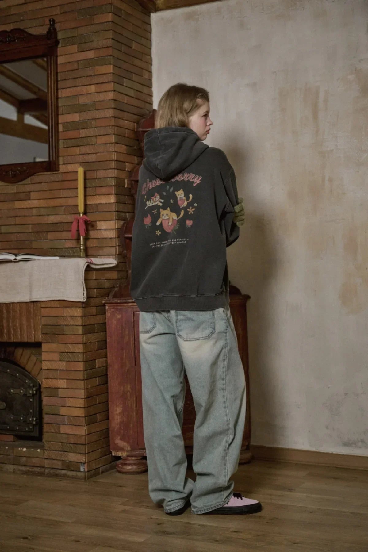 Woman wearing dark hooded zip-up sweatshirt with cat graphics, loose light blue jeans, and pink black sneakers indoors