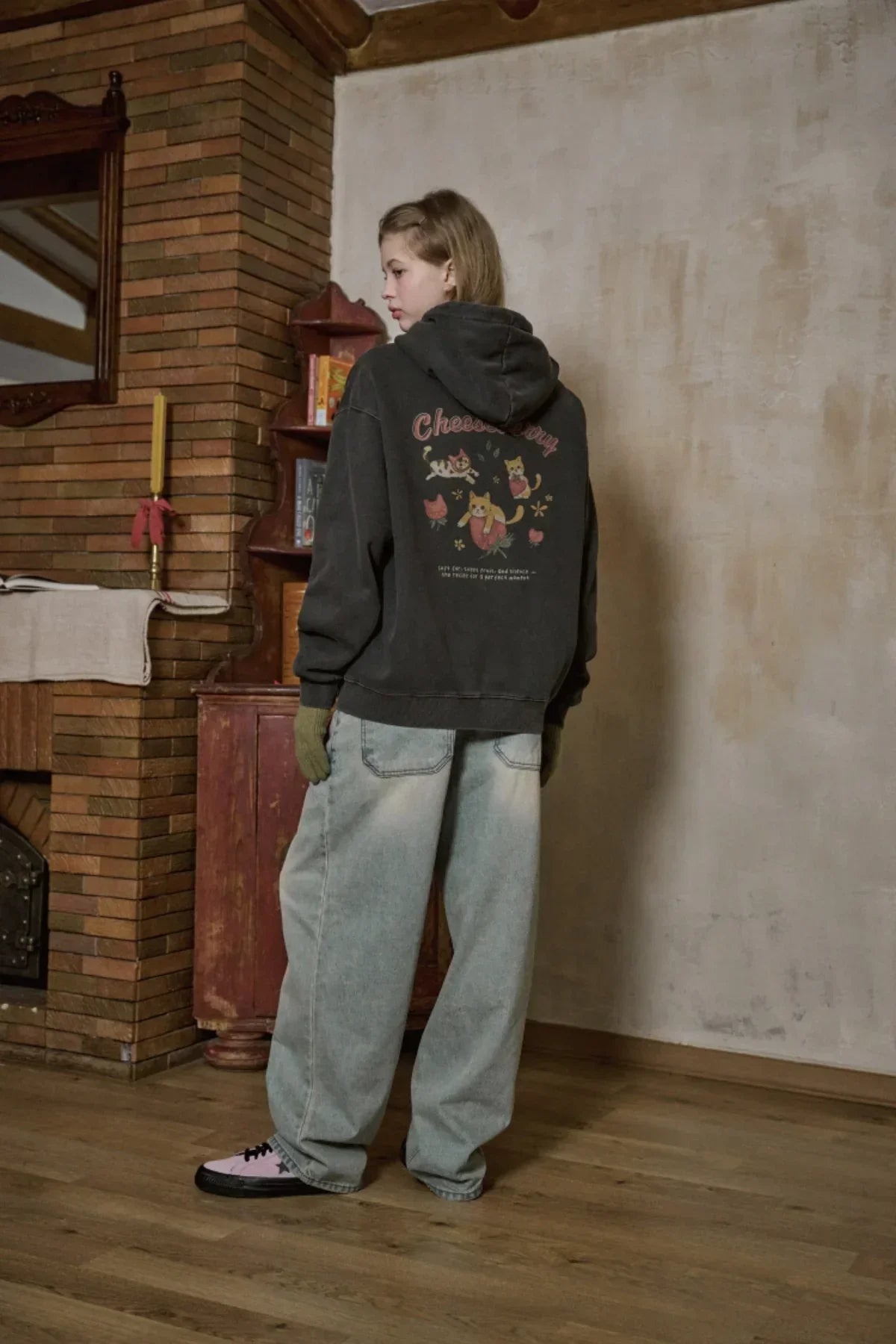 Young person indoors wearing black Cheeseberry graphic hoodie, wide light blue jeans, green gloves, and pink sneakers