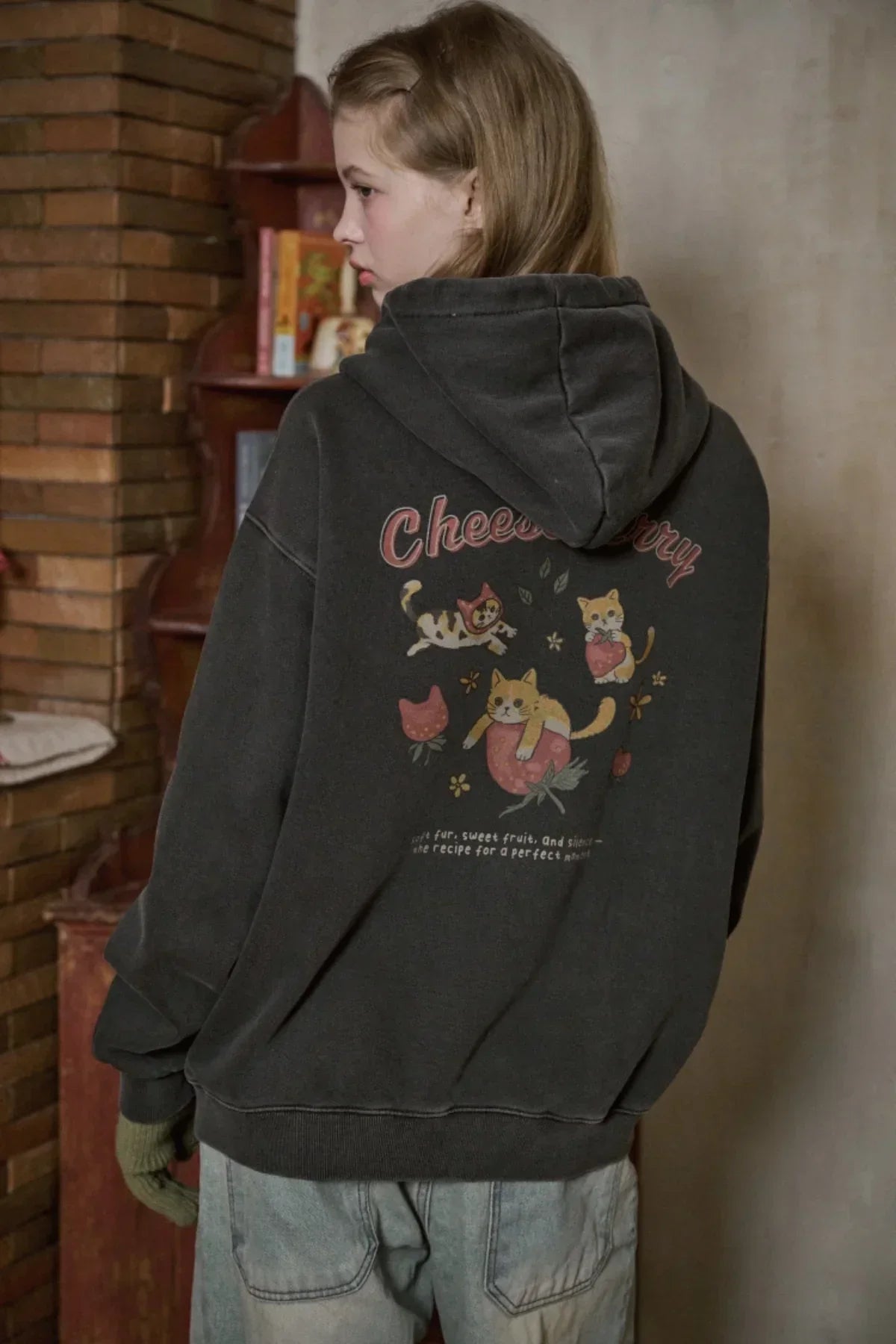 Woman wearing black hooded zip-up sweatshirt with cats and text Cheeberry in cozy indoor setting