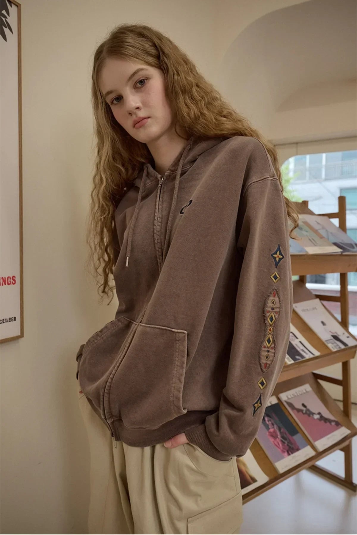 Young woman with long curly hair wearing brown zip-up hoodie with embroidered sleeve designs indoors