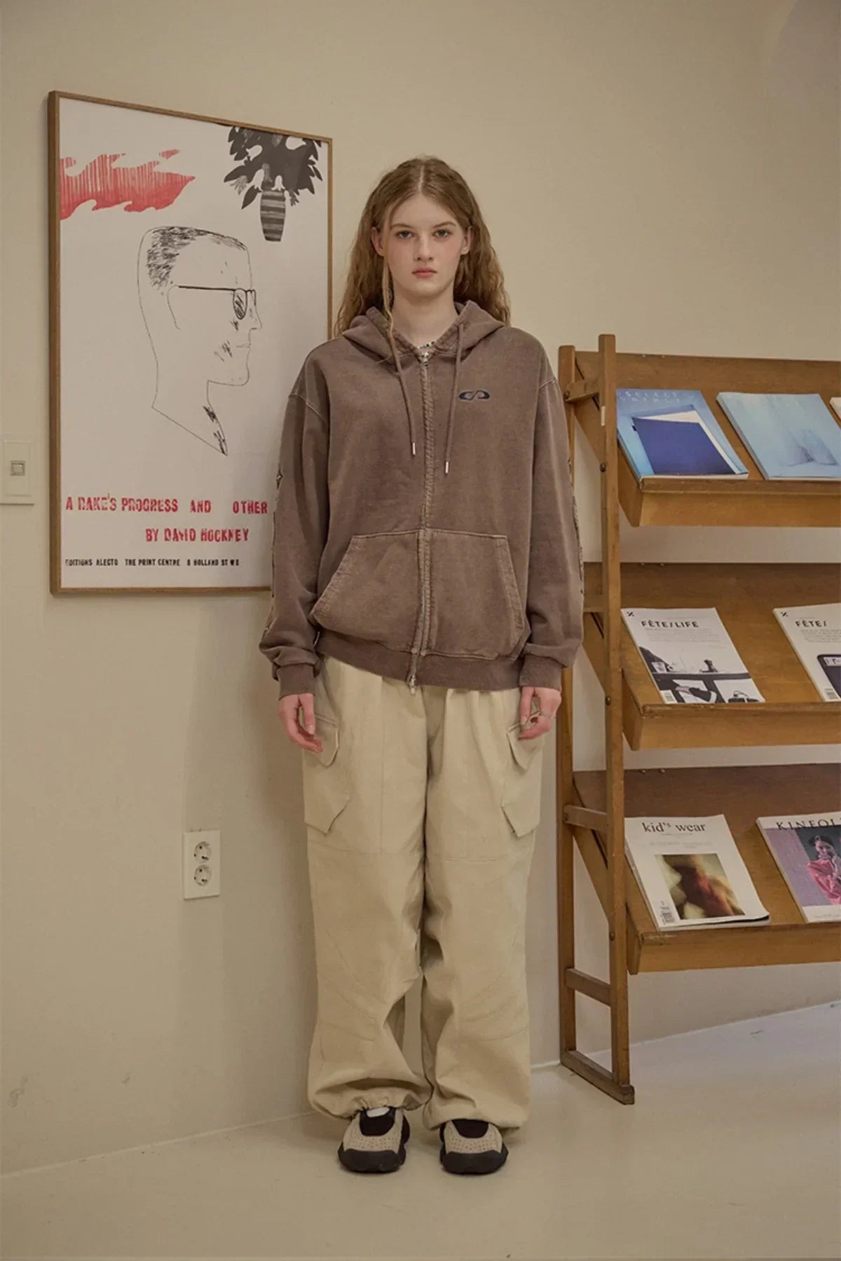 Young woman wearing brown zip-up hoodie and beige cargo pants standing indoors near poster and magazine rack