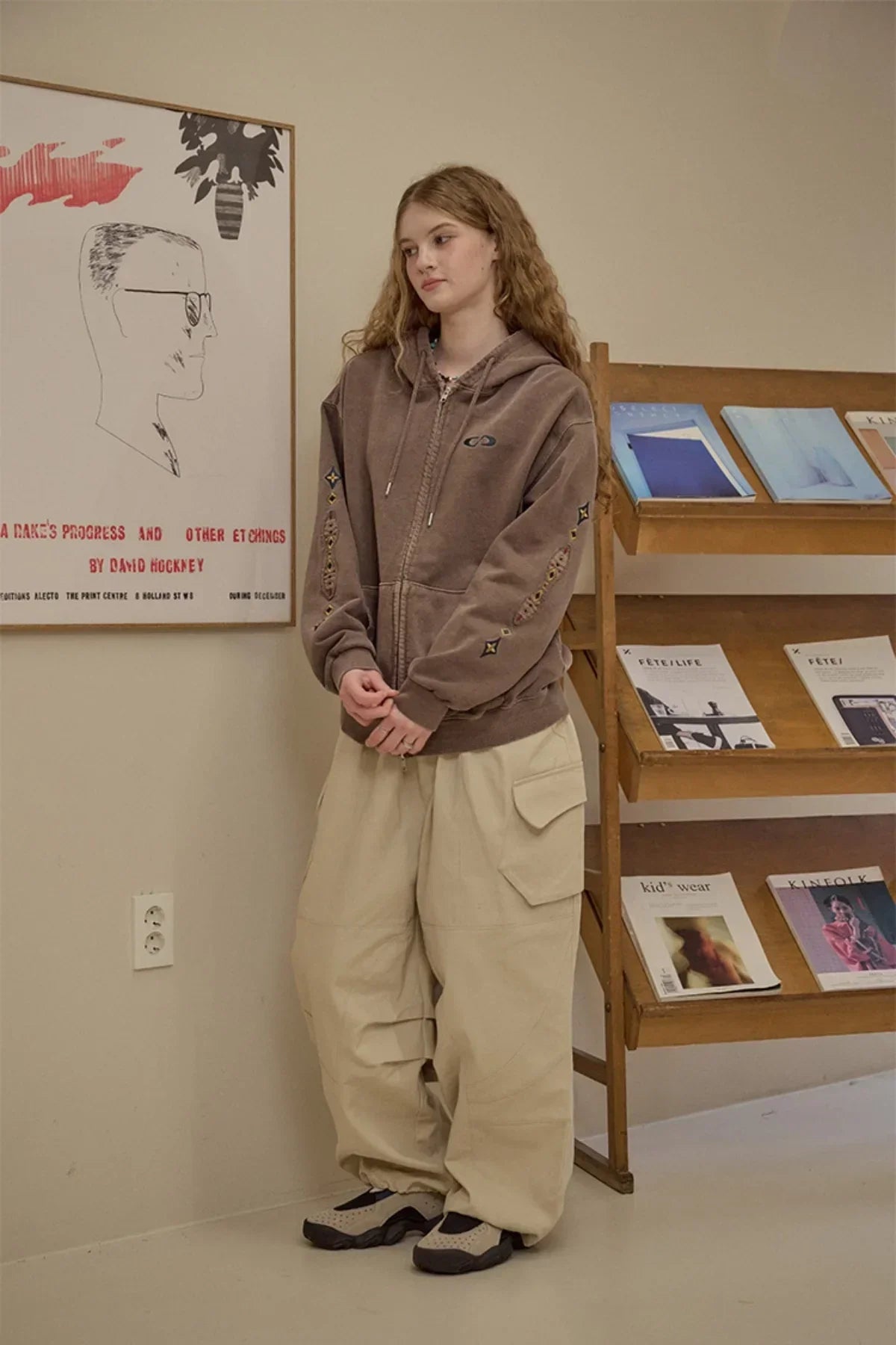 Young woman wearing brown zip-up hoodie with embroidered sleeves and beige cargo pants indoors
