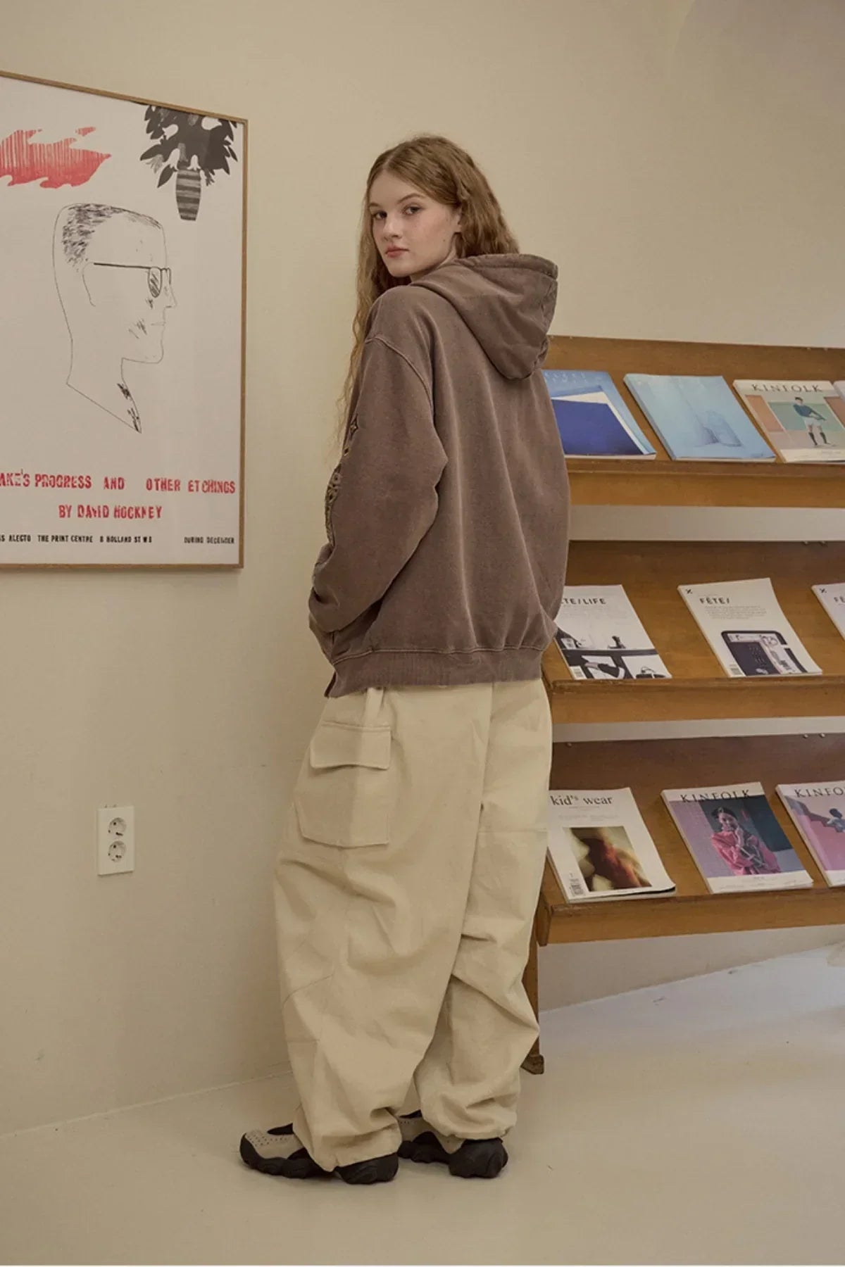 Young woman wearing brown hooded jacket and baggy beige cargo pants in minimalist room with art and magazines