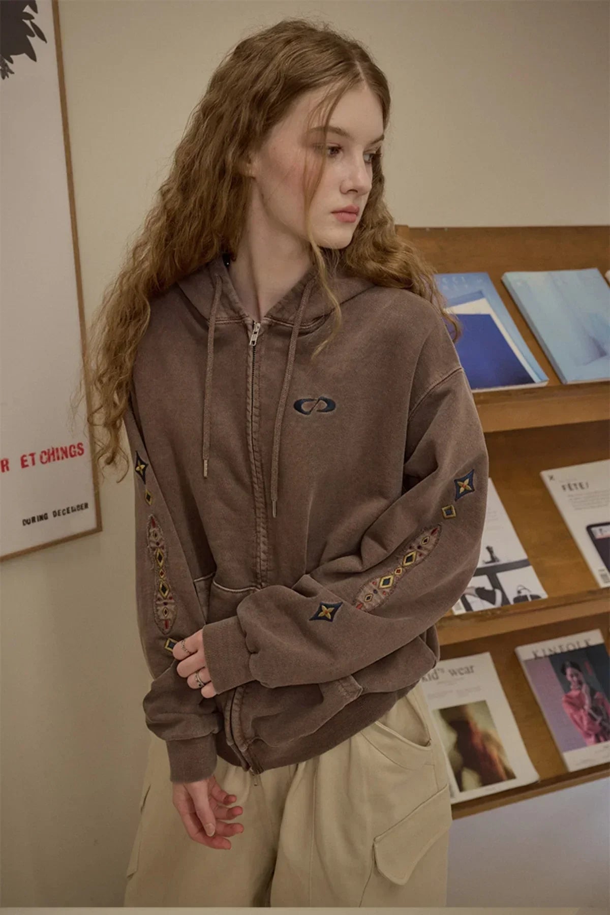 Young woman wearing brown zip-up hoodie with embroidered sleeves and beige pants inside a room