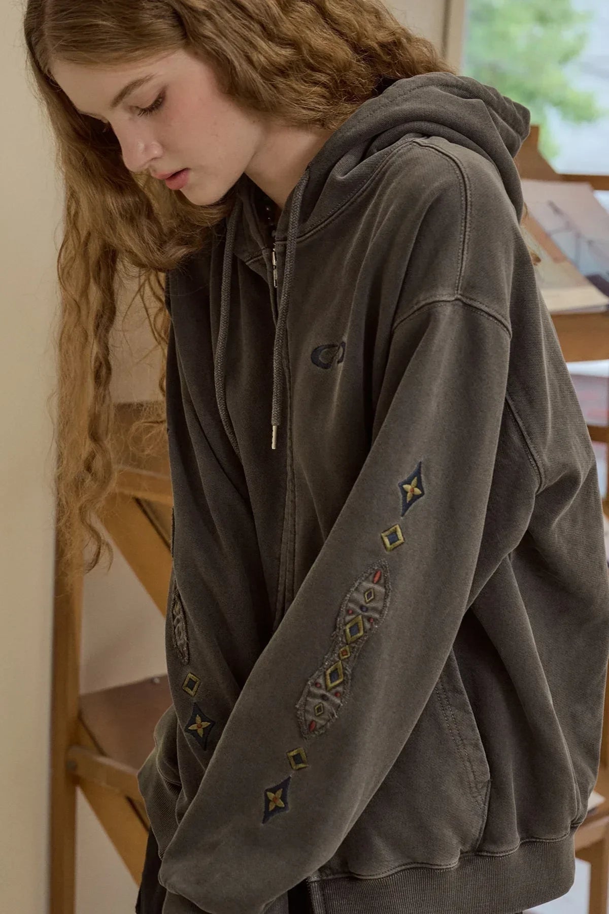 Close-up of woman wearing smoke black hooded zip-up with embroidered geometric patches on sleeves indoors