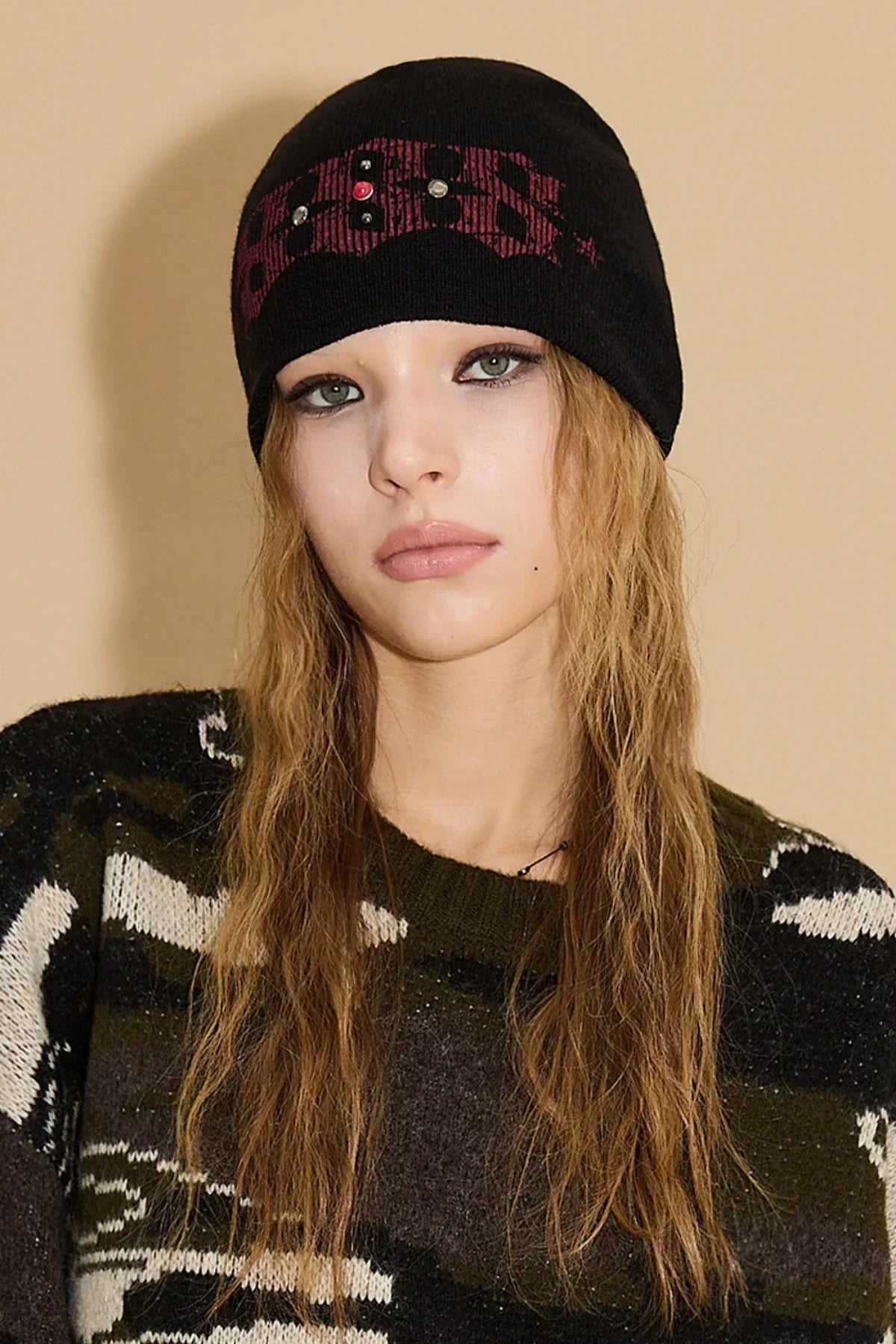 Young woman wearing a black studded beanie and patterned dark sweater against beige background