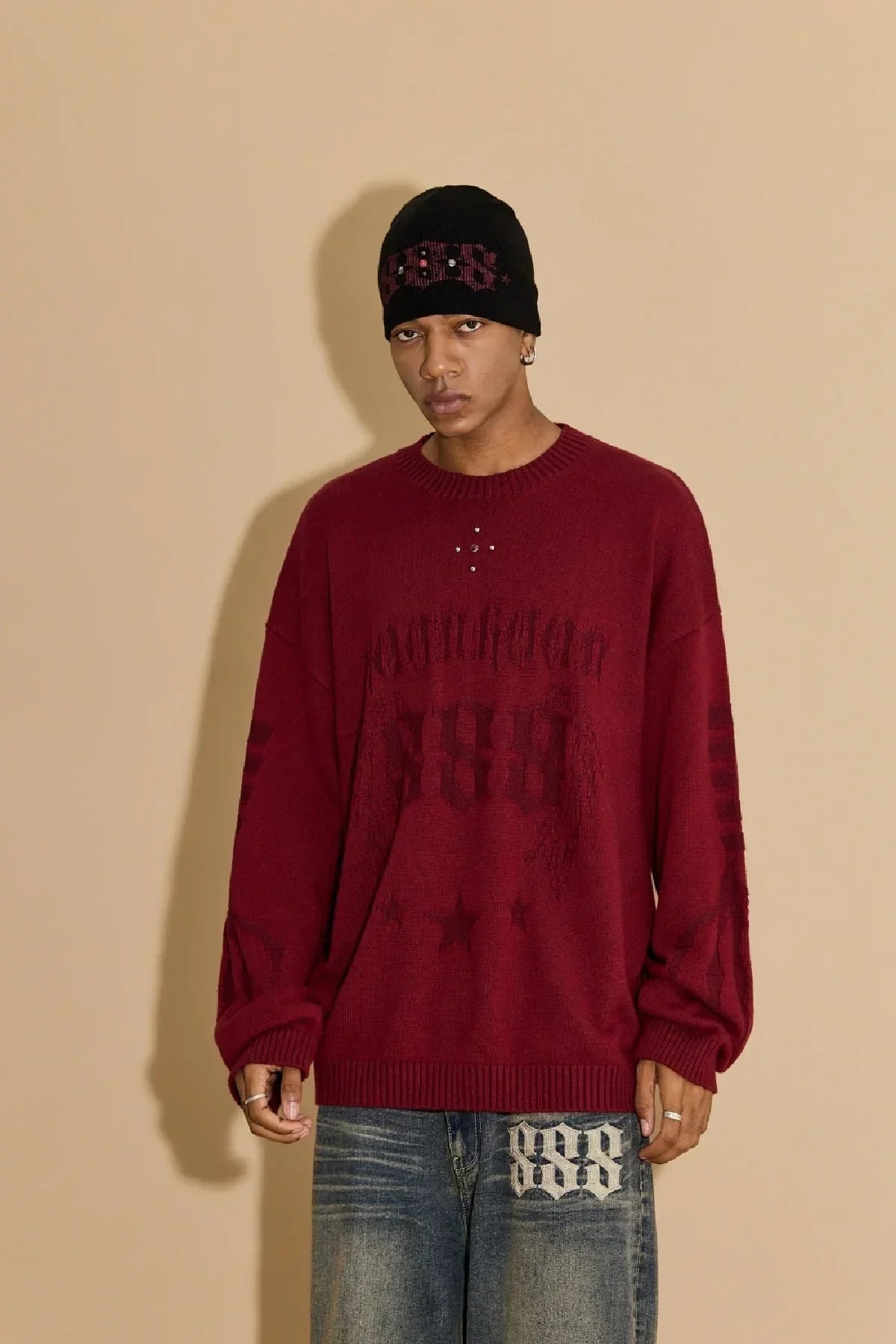 Male model wearing black studded beanie, red oversized sweater with graphic print, and faded jeans against beige background