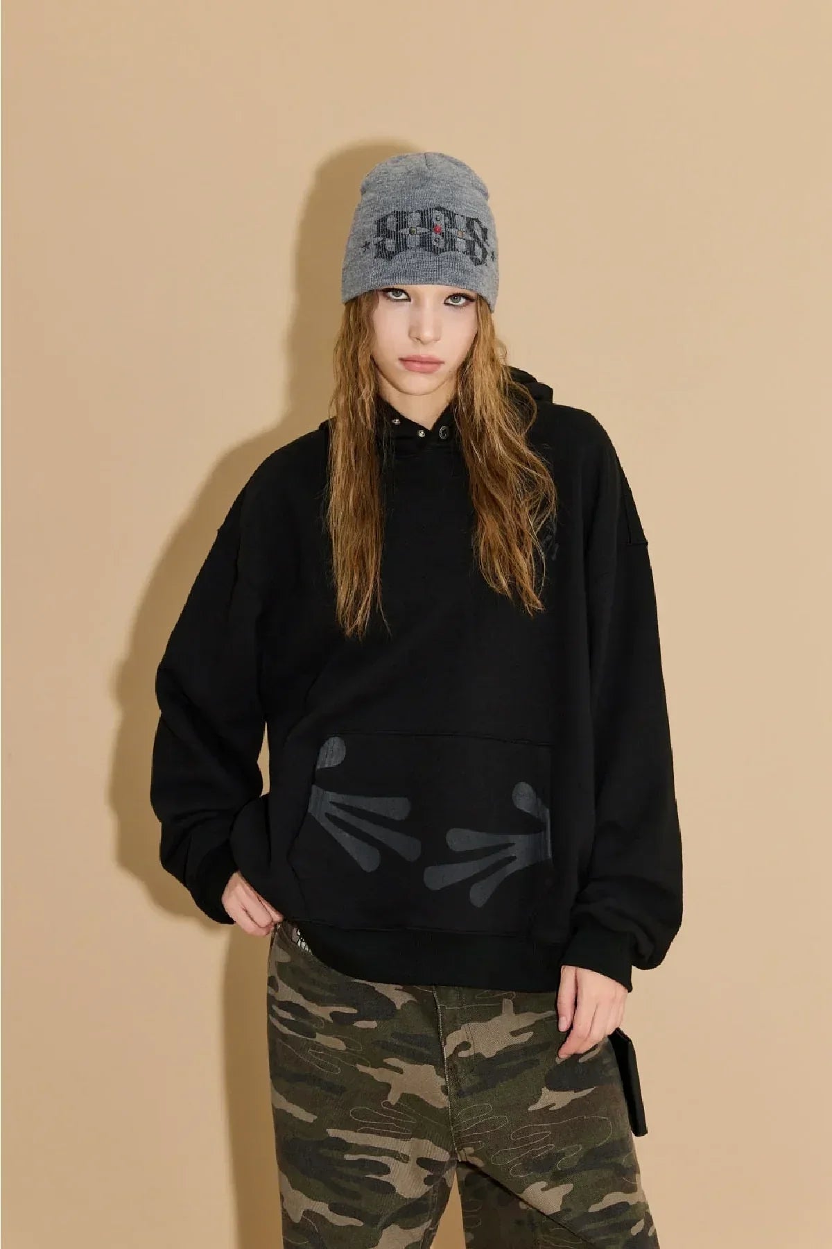 Young woman wearing grey studded beanie, black hoodie with handprint design, and camo pants