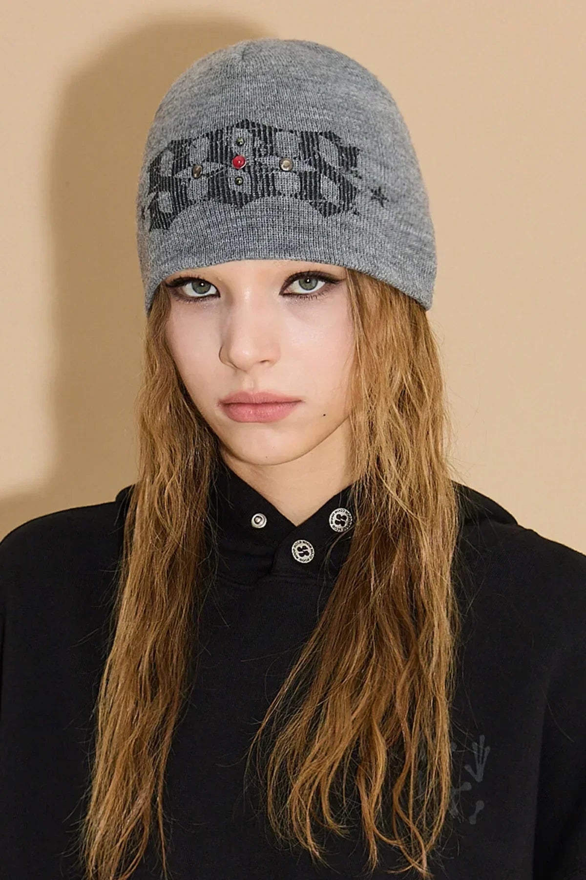 Close-up of a woman with long wavy hair wearing a grey studded beanie and black sweatshirt against beige background