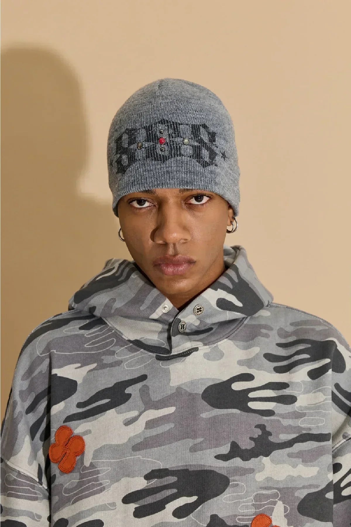 Portrait of person wearing grey studded graphic beanie and camo hoodie with orange patch