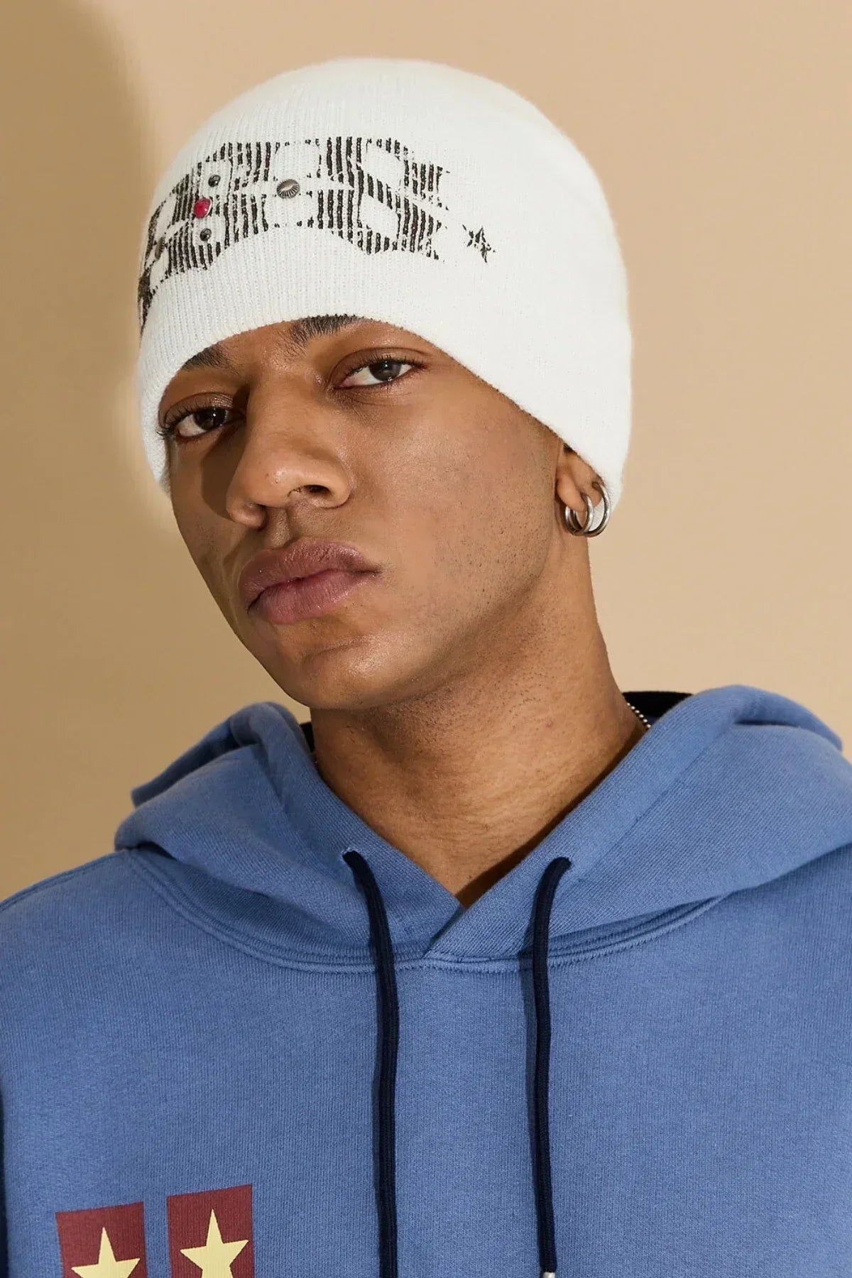 Close-up of man wearing ivory studded beanie with text and blue hoodie against beige background