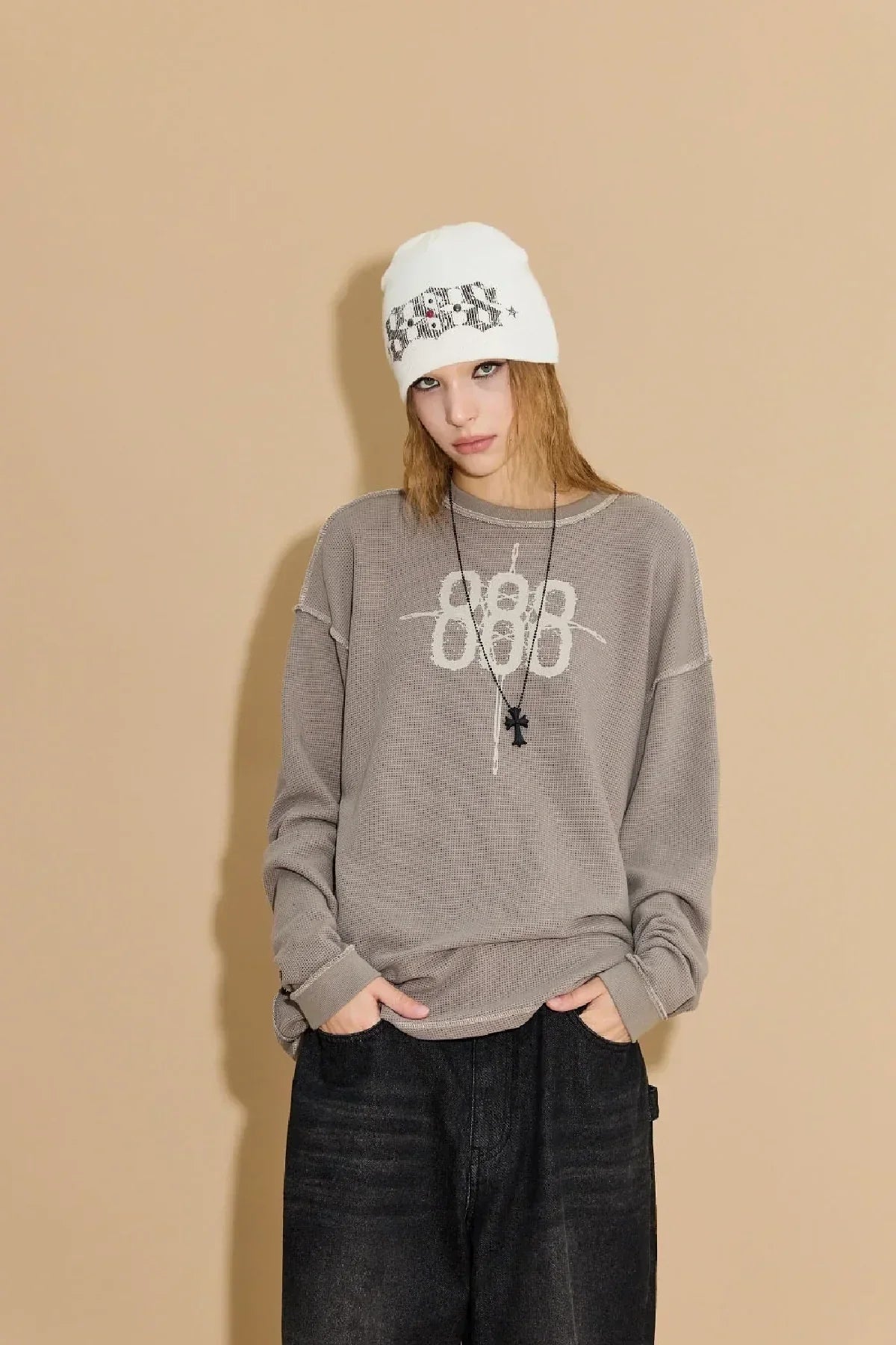 Model wearing ivory 888 stud beanie, beige 888 logo sweater, black pants, and black cross necklace