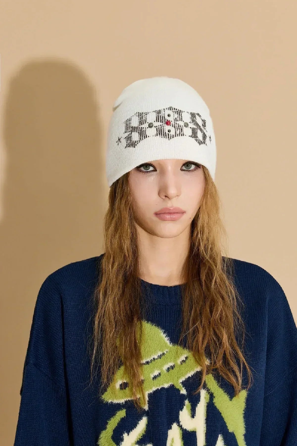 Young woman wearing an ivory knit beanie with studded logo and navy blue sweater with green UFO graphic