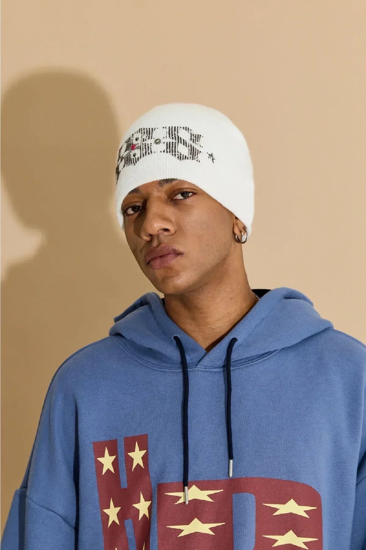 Young man wearing ivory studded beanie and blue hoodie with star print against beige background