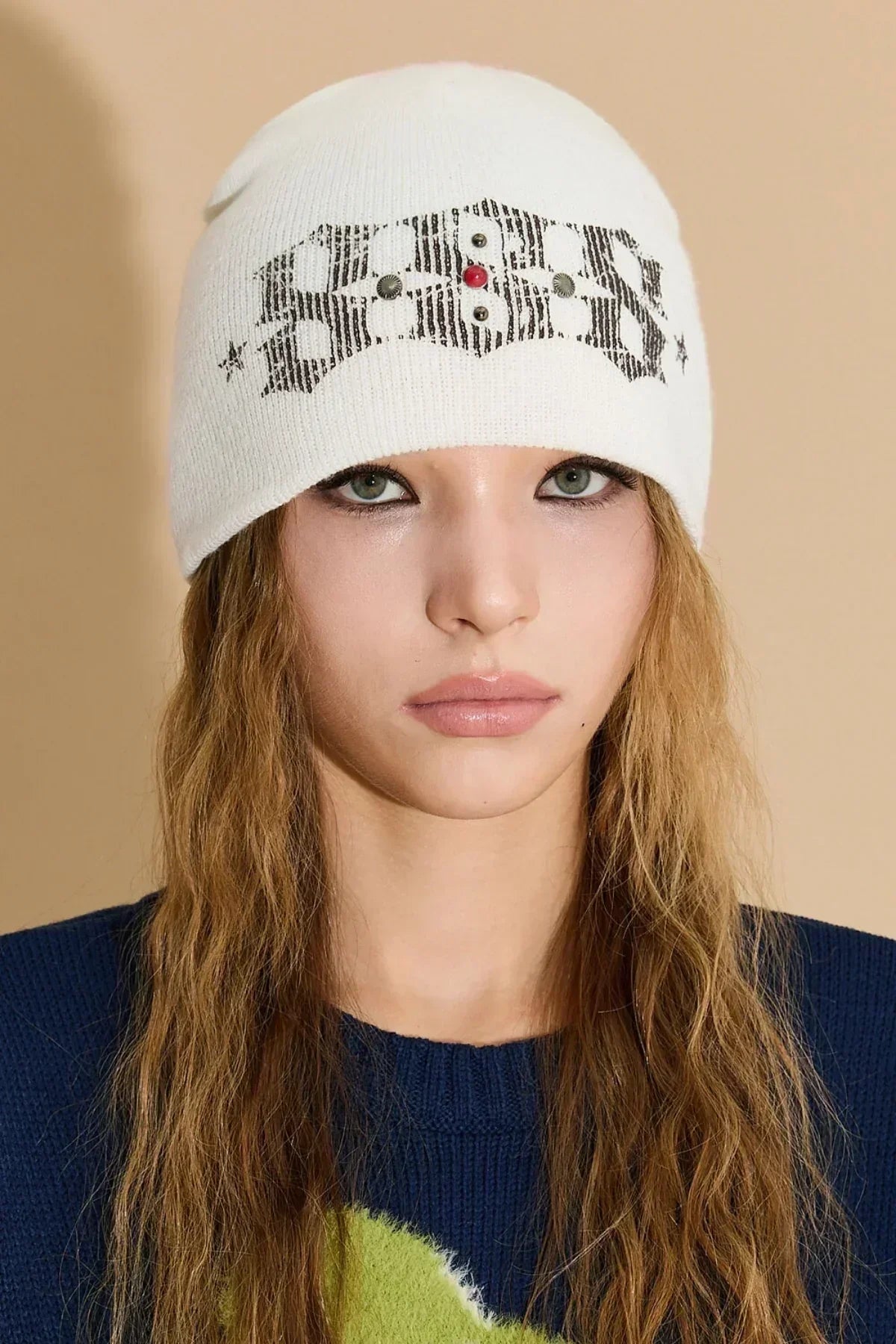 Young woman with wavy hair wearing a white studded beanie hat and navy sweater in studio