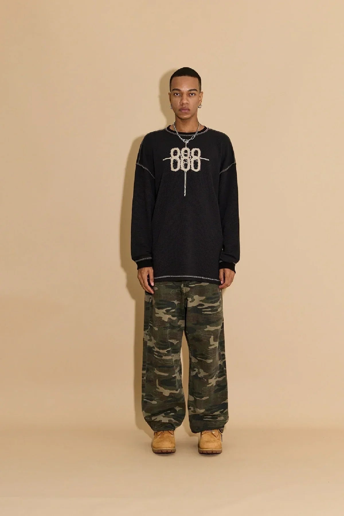 Man wearing black 888 waffle long sleeve T-shirt with camo cargo pants and tan boots on beige background