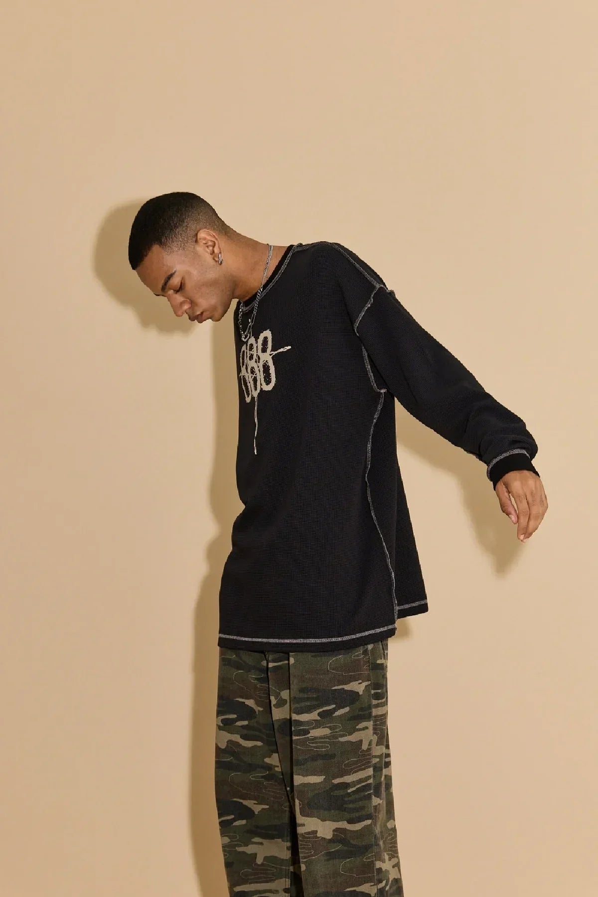 Male model wearing black 888 waffle long sleeve t-shirt with camo pants against beige background
