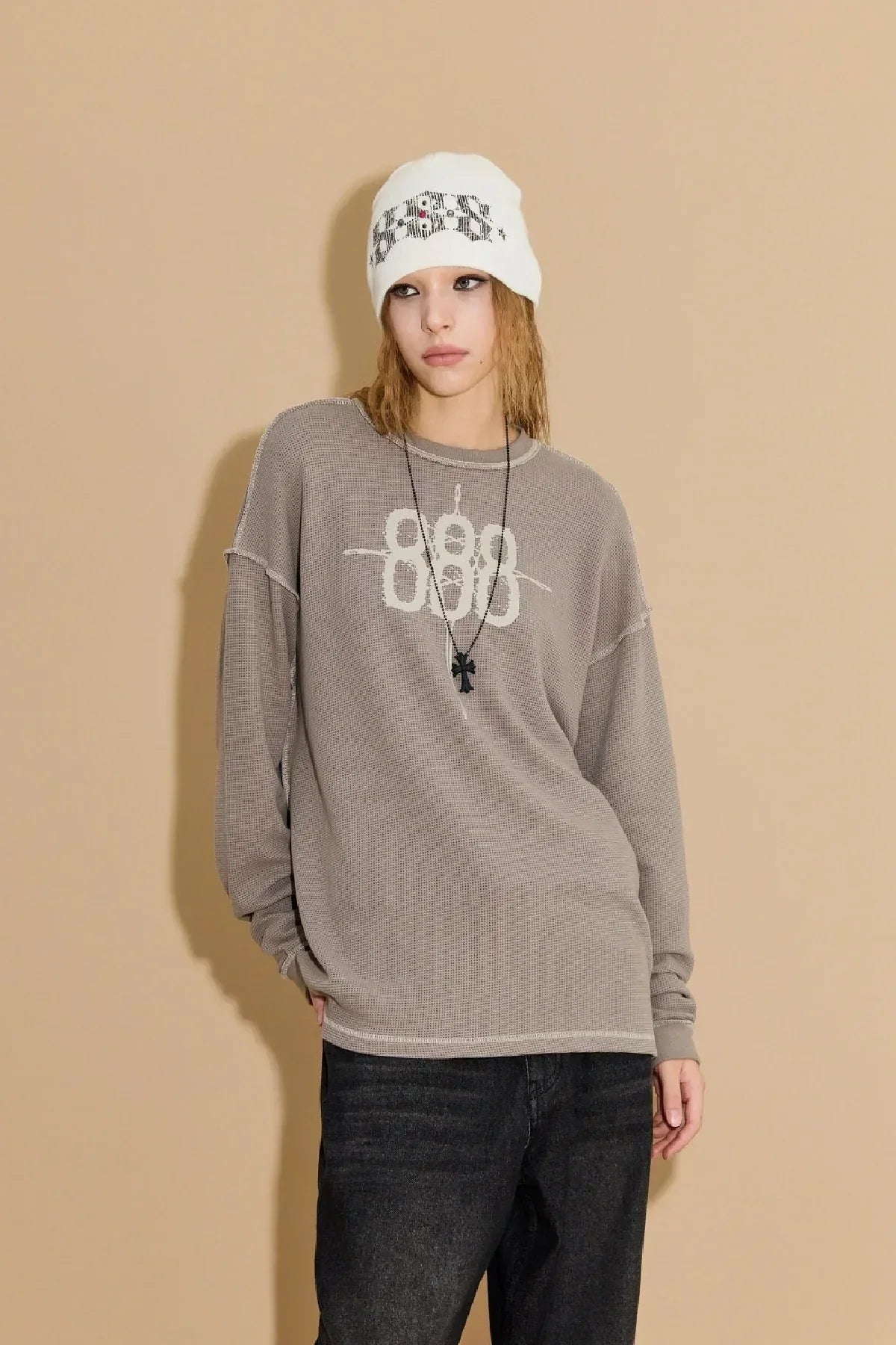 Model wearing greyish beige waffle long sleeve t-shirt with white 888 graphic and black cross necklace, white beanie