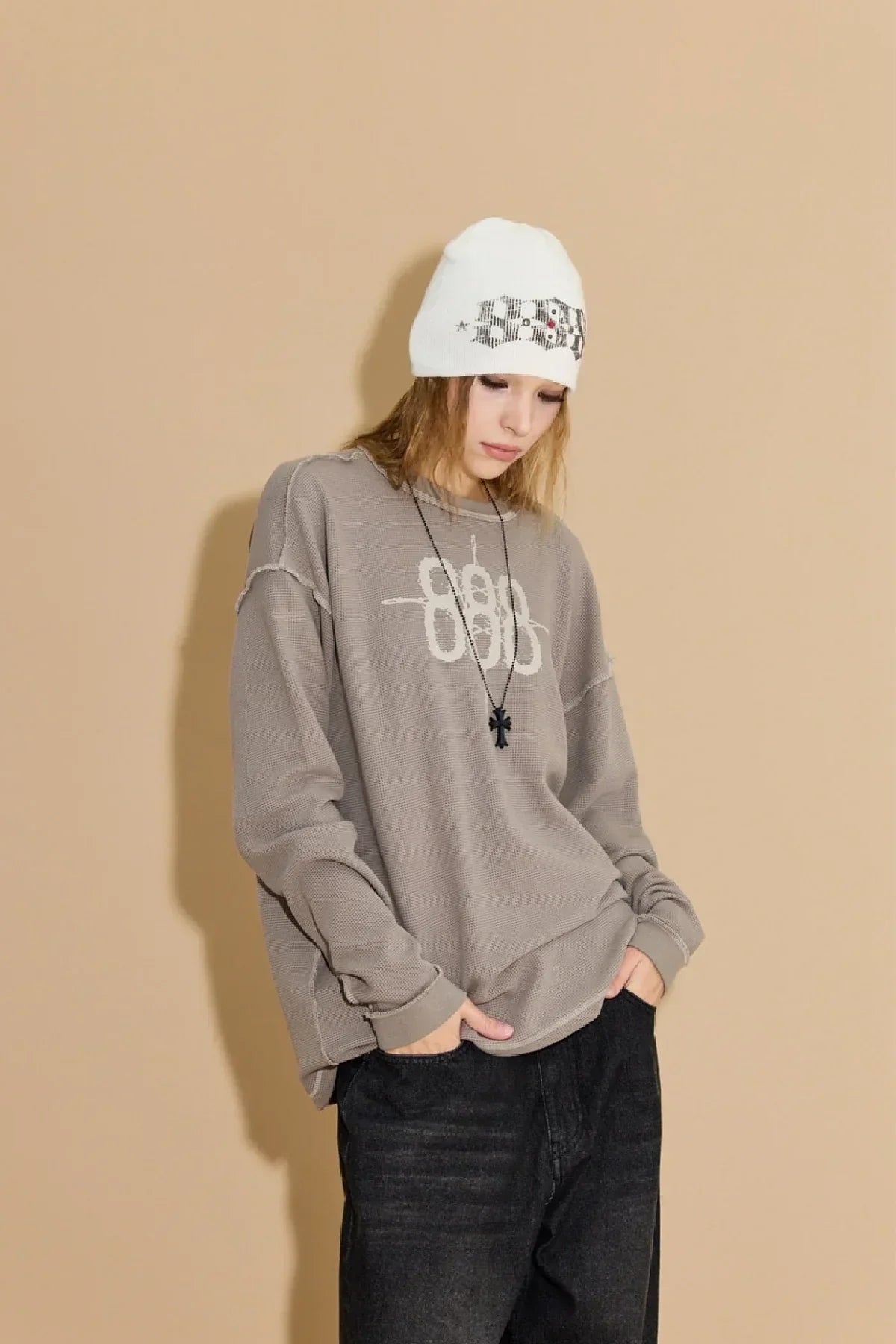Young woman wearing greyish beige waffle long sleeve shirt and white beanie, posing against beige background