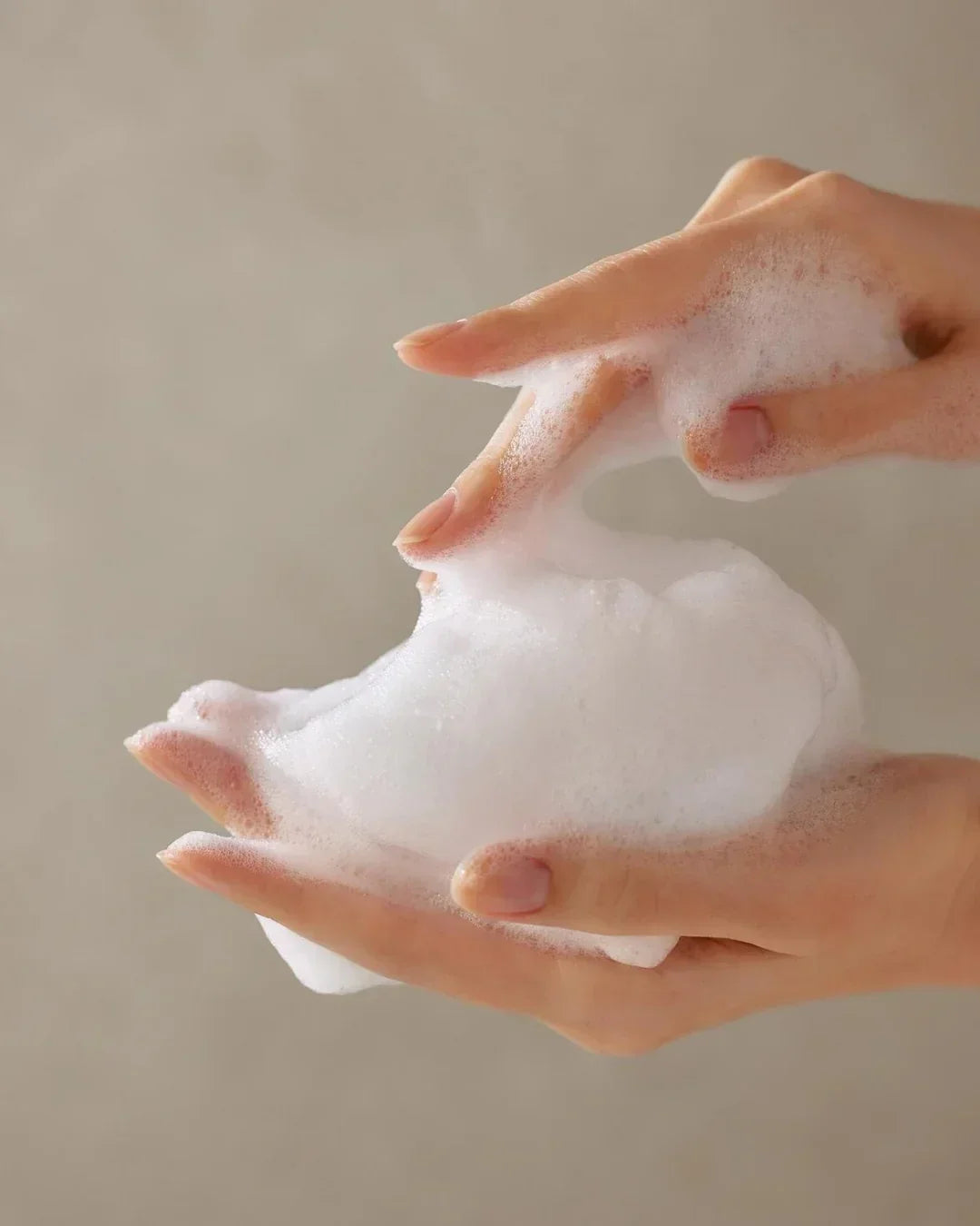 Hands holding and playing with rich white foamy cleanser foam against neutral background