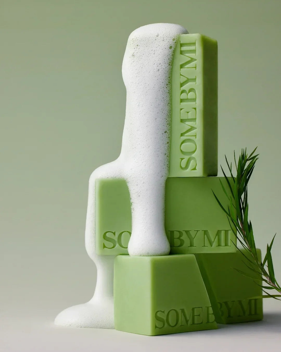 Green SOMEBYMI soap bars stacked with white foam and green plant branch on light green background