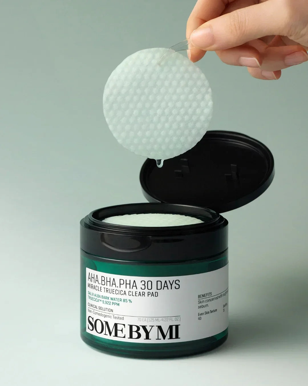 Hand holding AHA BHA PHA 30 Days Miracle Truecica Clear Pad with droplet over open black green container