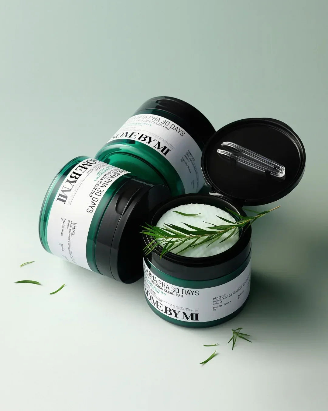 Two green jars of SOME BY MI AHA BHA PHA 30 Days Miracle Clear Pad with cotton pads and sprigs on pale green surface