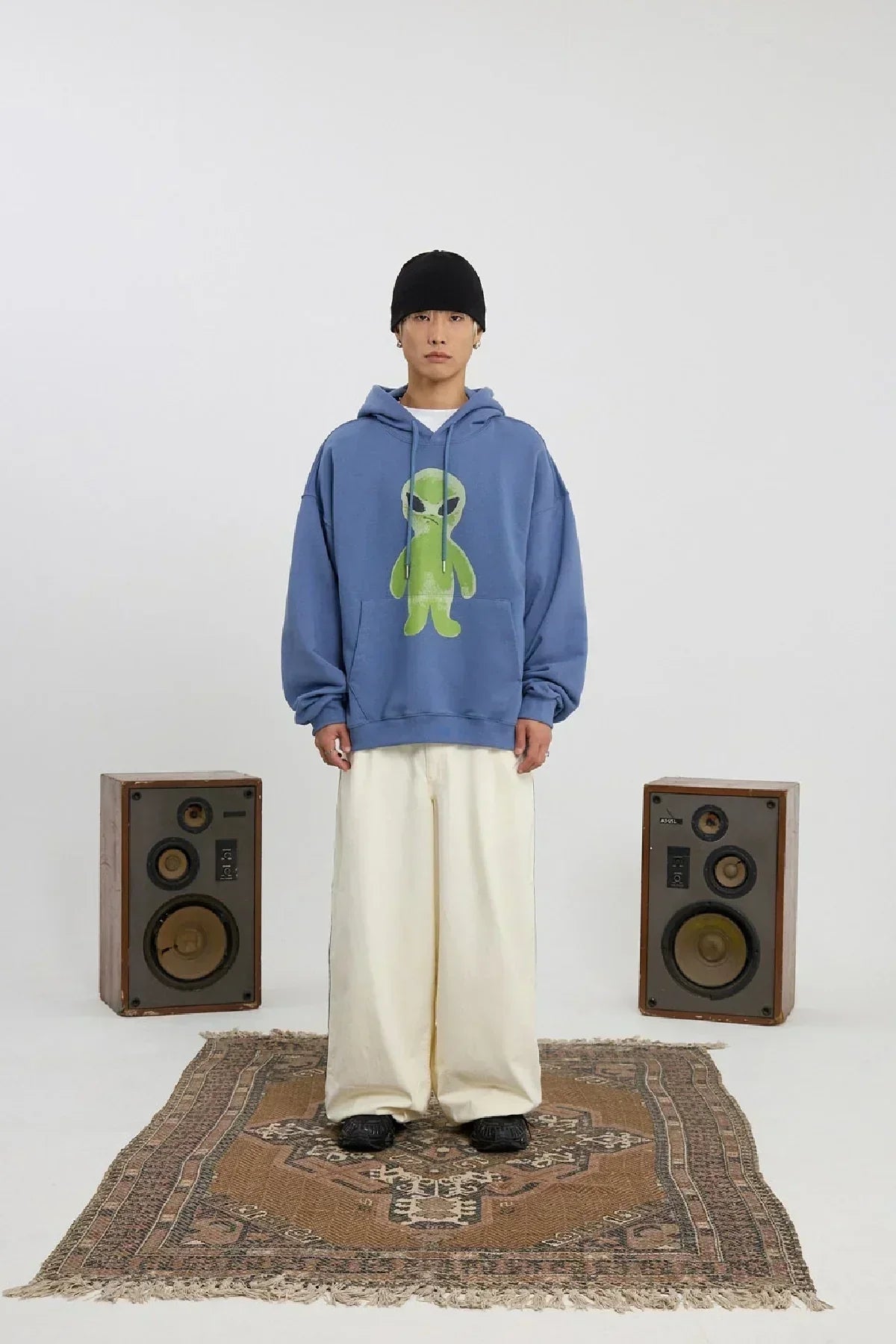 Man wearing dust blue hoodie with green alien graphic, wide cream pants, black beanie, standing on patterned rug