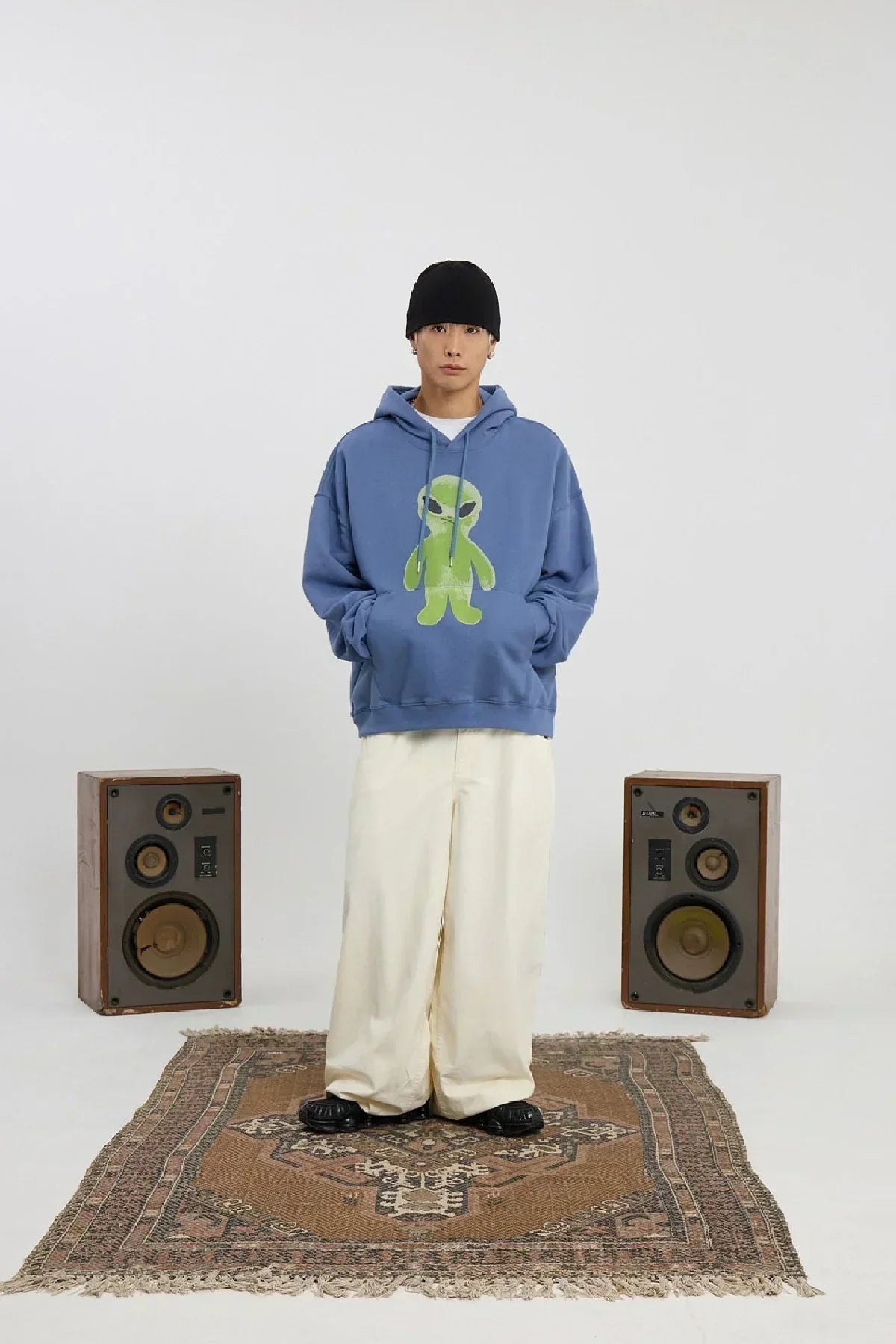 Man wearing dust blue hoodie with green alien graphic, white wide pants, black beanie, standing on patterned rug