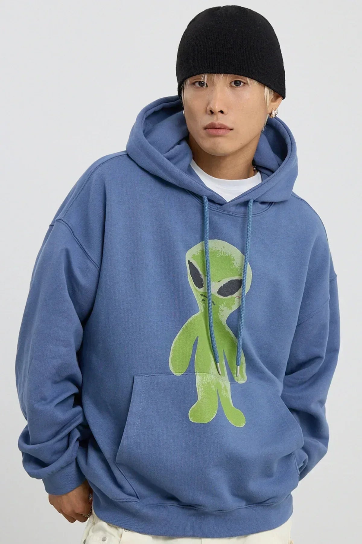 Man wearing dust blue hoodie with green alien graphic, black beanie, and white pants in studio