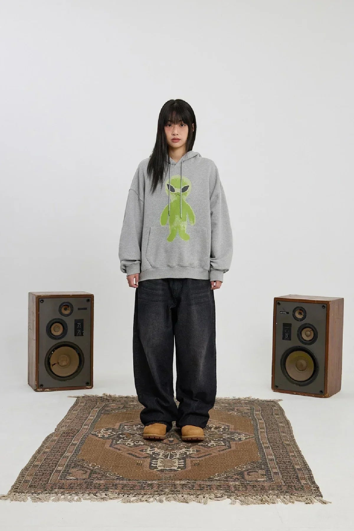 Young woman wearing grey hoodie with green alien graphic, wide black pants, and brown shoes, standing on patterned rug