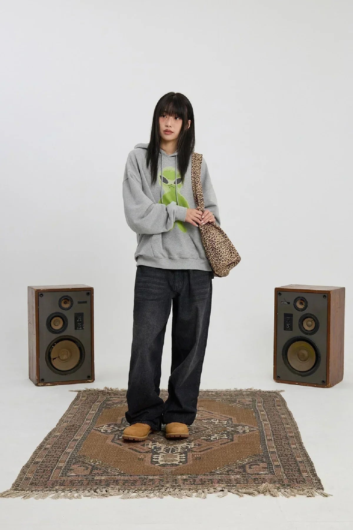 Woman wearing grey alien graphic hoodie, black wide pants, tan boots, and leopard print shoulder bag on patterned rug with vintage speakers