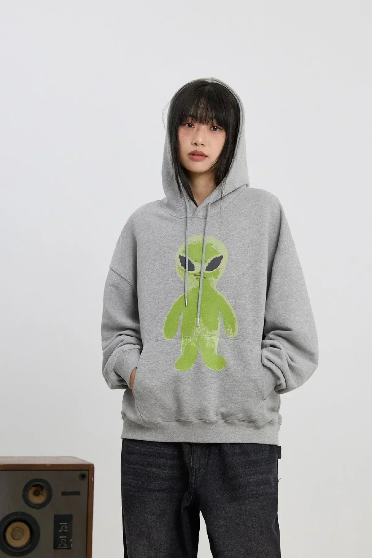 Young woman wearing gray hoodie with green alien graphic standing against plain background