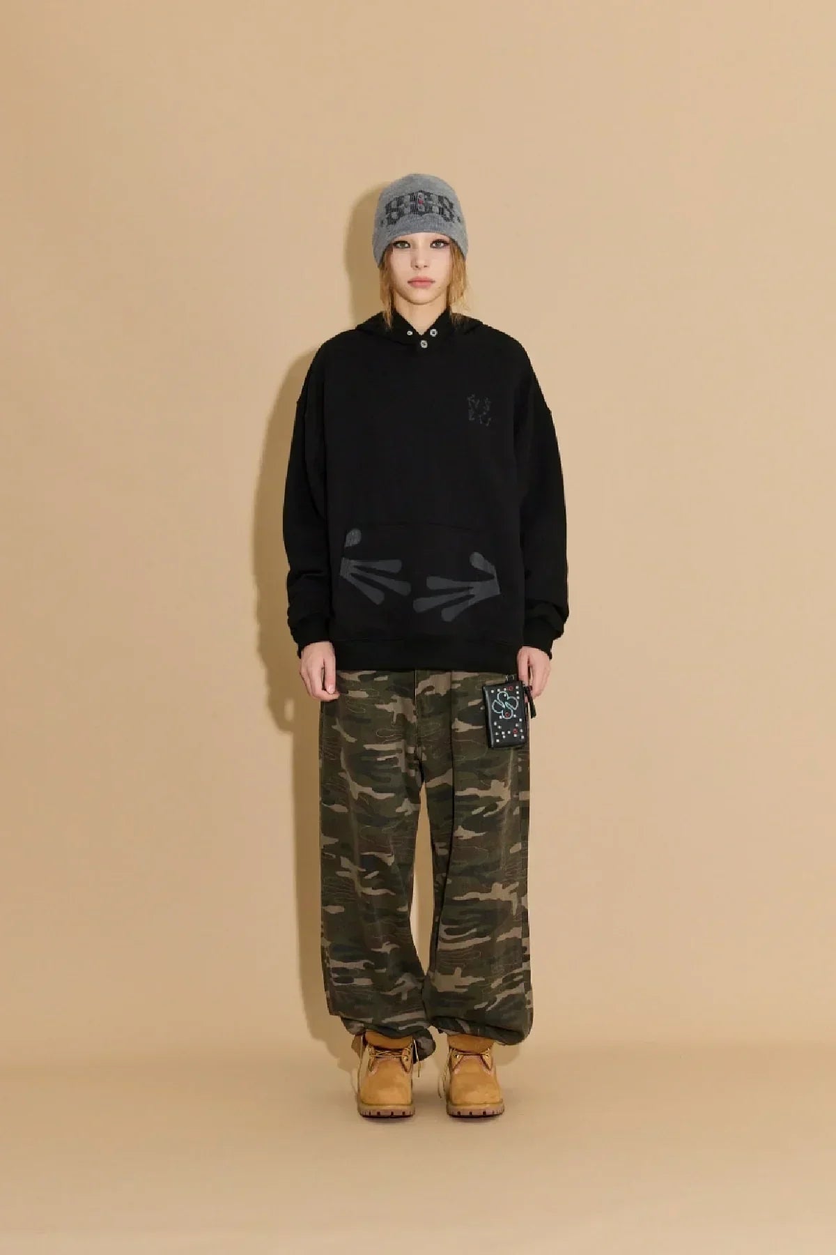 Model wearing black hoodie with alien hands print, camo pants, grey beanie, and tan boots
