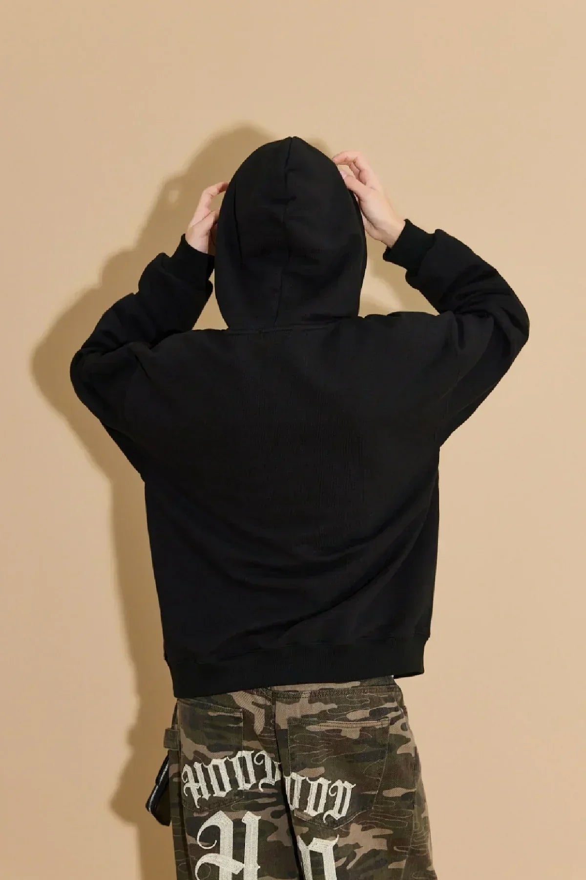 Person wearing black hoodie with hood up and camouflage pants with white graffiti writing