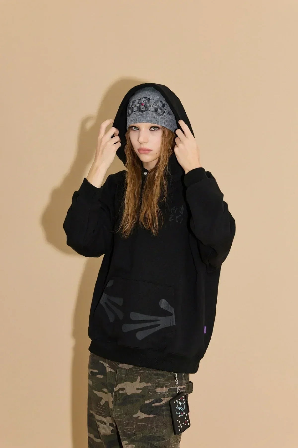 Young woman wearing black hoodie with alien hand prints and gray beanie, camouflage pants, beige background